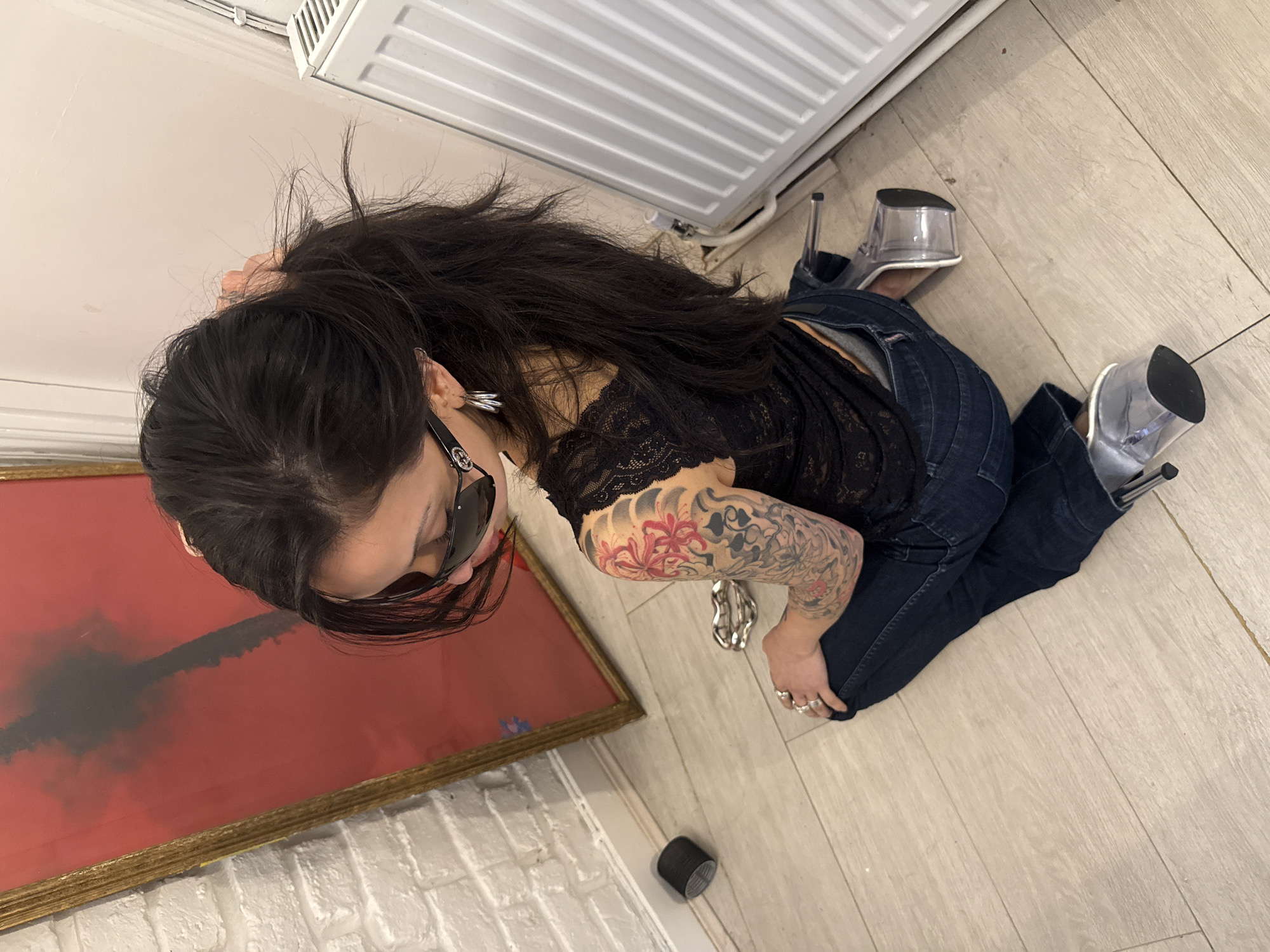 A woman with long dark hair, wearing sunglasses, a black lace top, dark jeans, and clear platform heels, is kneeling on a light wood-grain floor. Her right arm, which has a floral tattoo, is resting on the floor near a framed red painting with a black streak in the center. There is a white radiator on the wall behind her and a black object on the floor near a white brick wall in the lower left. The location is indoors. There are no indicators of the city of London, United Kingdom.
