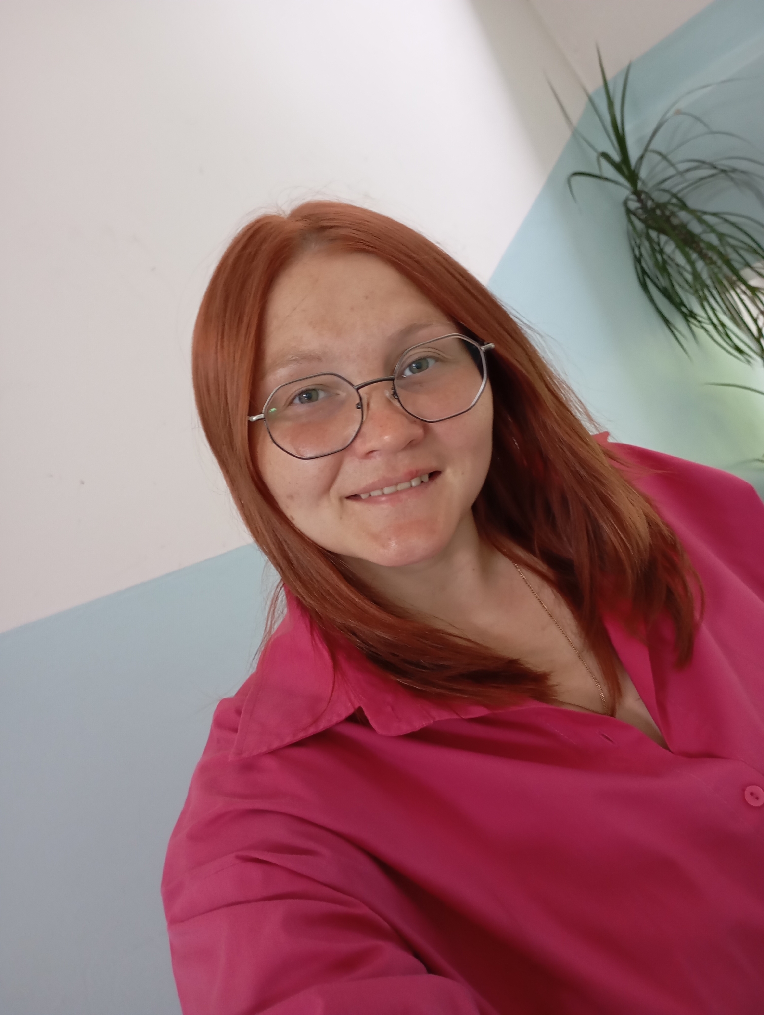 A woman with reddish-brown hair and round, dark-framed glasses is captured in a close-up, chest-up shot. She is smiling gently at the camera, with a friendly expression. Her eyes are light-colored, and some freckles are visible on her face. She is wearing a bright, fuchsia-pink button-up shirt with a collar. A thin, gold-colored necklace is partially visible around her neck.
The background features an indoor setting with a wall painted in two distinct sections: white on the upper part and a light, pastel blue on the lower part, creating a clean, modern aesthetic. In the upper right corner, a plant with long, thin green leaves, possibly a Dracaena, adds a touch of greenery to the scene. The lighting appears bright and even, suggesting a well-lit interior, likely during the daytime. The overall mood of the image is positive and approachable. No text is visible in the image.