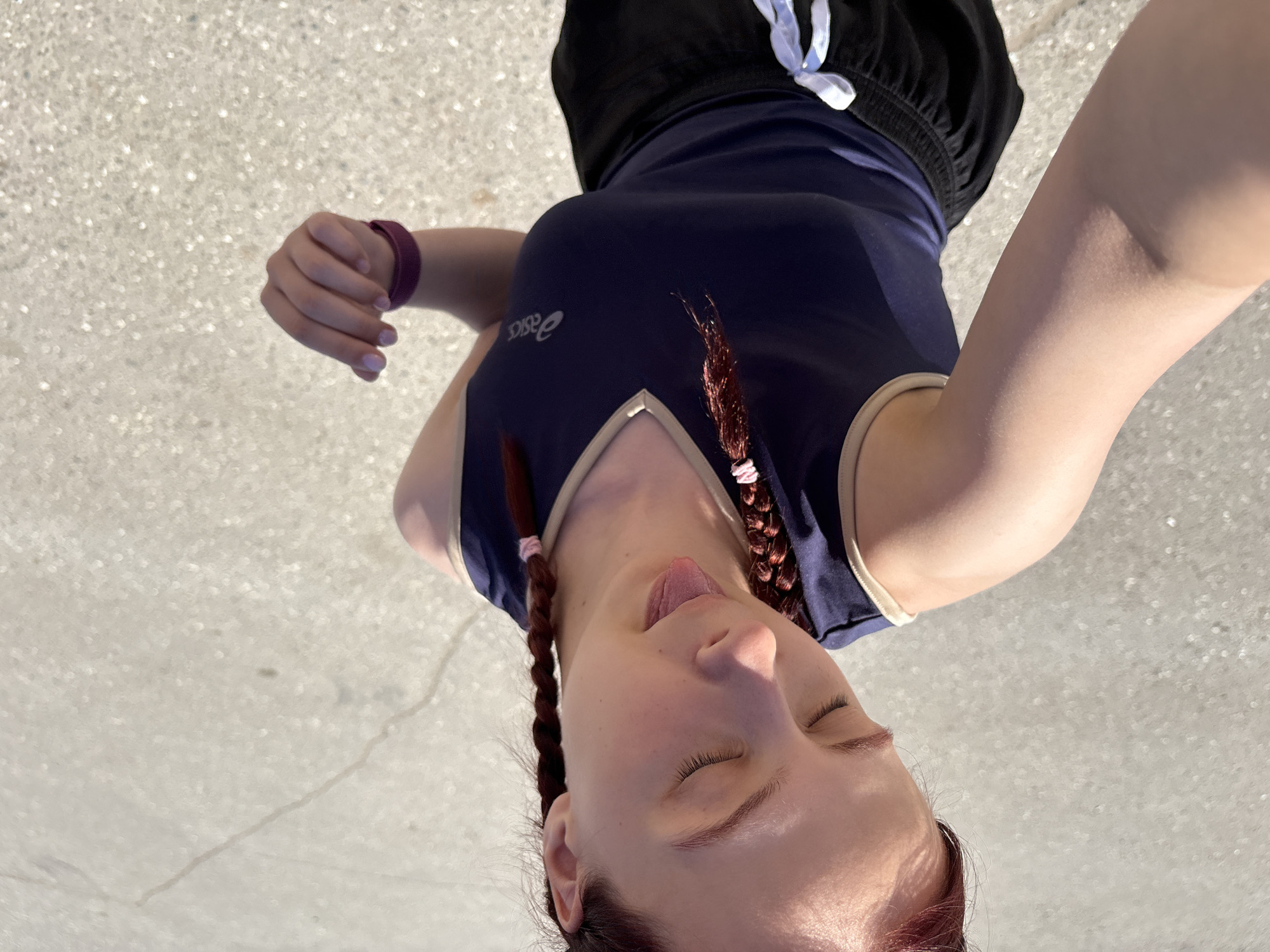 A high-angle shot depicts a woman with red hair styled in pigtails, wearing a blue athletic top with the word "Asics" visible on the left side. She has her eyes closed and tongue slightly protruding. She is also wearing a burgundy fitness tracker. The background consists of a concrete surface. There are no explicit indicators to confirm the location as Grishanki, Russia.
