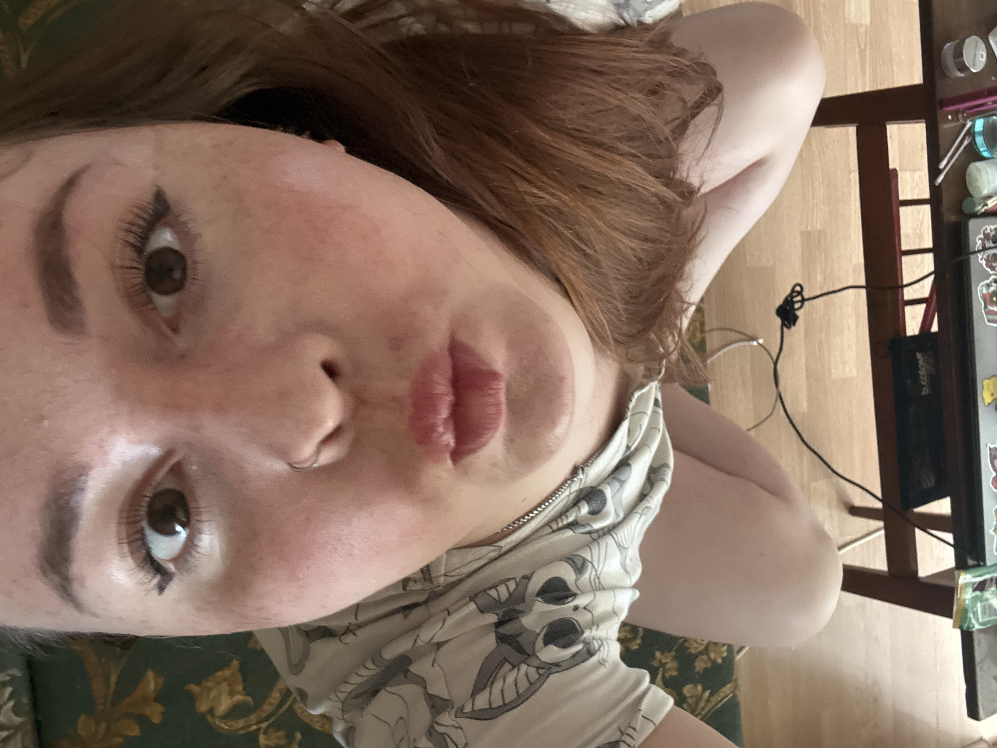 A selfie of a person with fair skin and auburn hair. They are looking up at the camera with brown eyes and a nose ring. Their lips are slightly pursed. They are wearing a light-colored top with a repeating pattern. A chair and part of a desk are visible in the background, along with various objects on the desk. The floor is made of wooden planks. The location is not discernible from the image.