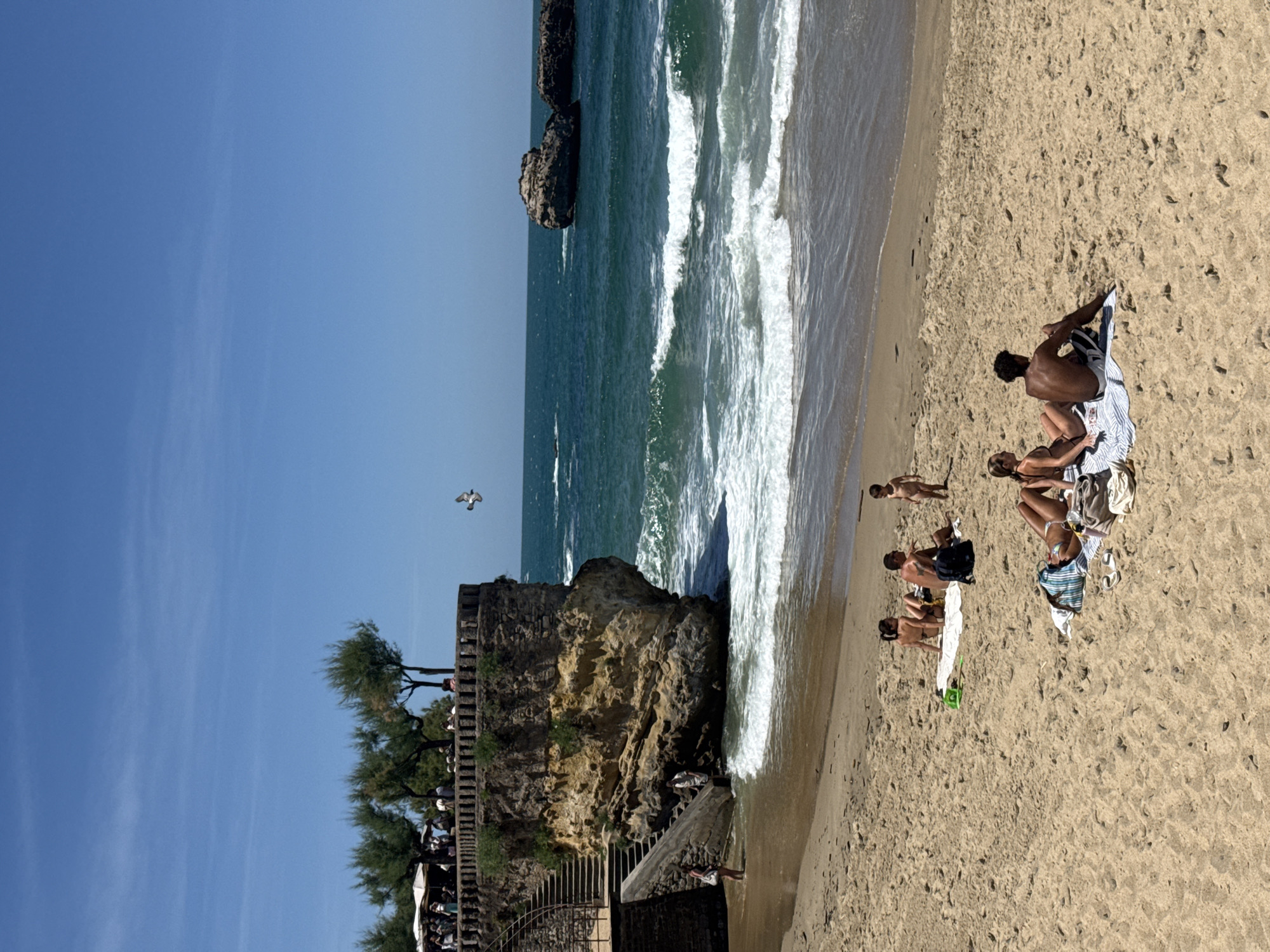 This is a photograph taken at the Rocher de la Vierge in Biarritz, France, on a sunny day. The scene depicts a beach with several people sunbathing and relaxing on the sand. In the foreground, two couples are spread out on towels. One couple, a man and a woman, are lying down, while the other couple, also a man and a woman, are sitting. Further up the beach, another group of people are gathered on towels, including a man, a woman, and a young child standing.
The background features the ocean with rolling waves and rocky outcrops in the distance. A prominent rocky cliff face juts out from the coastline, topped with trees and a pathway lined with people. Stone stairs descend along the cliff face, leading to the water's edge where a man and a woman are interacting. A bird is captured in flight against the clear blue sky. The sand on the beach is light brown and appears somewhat dry.