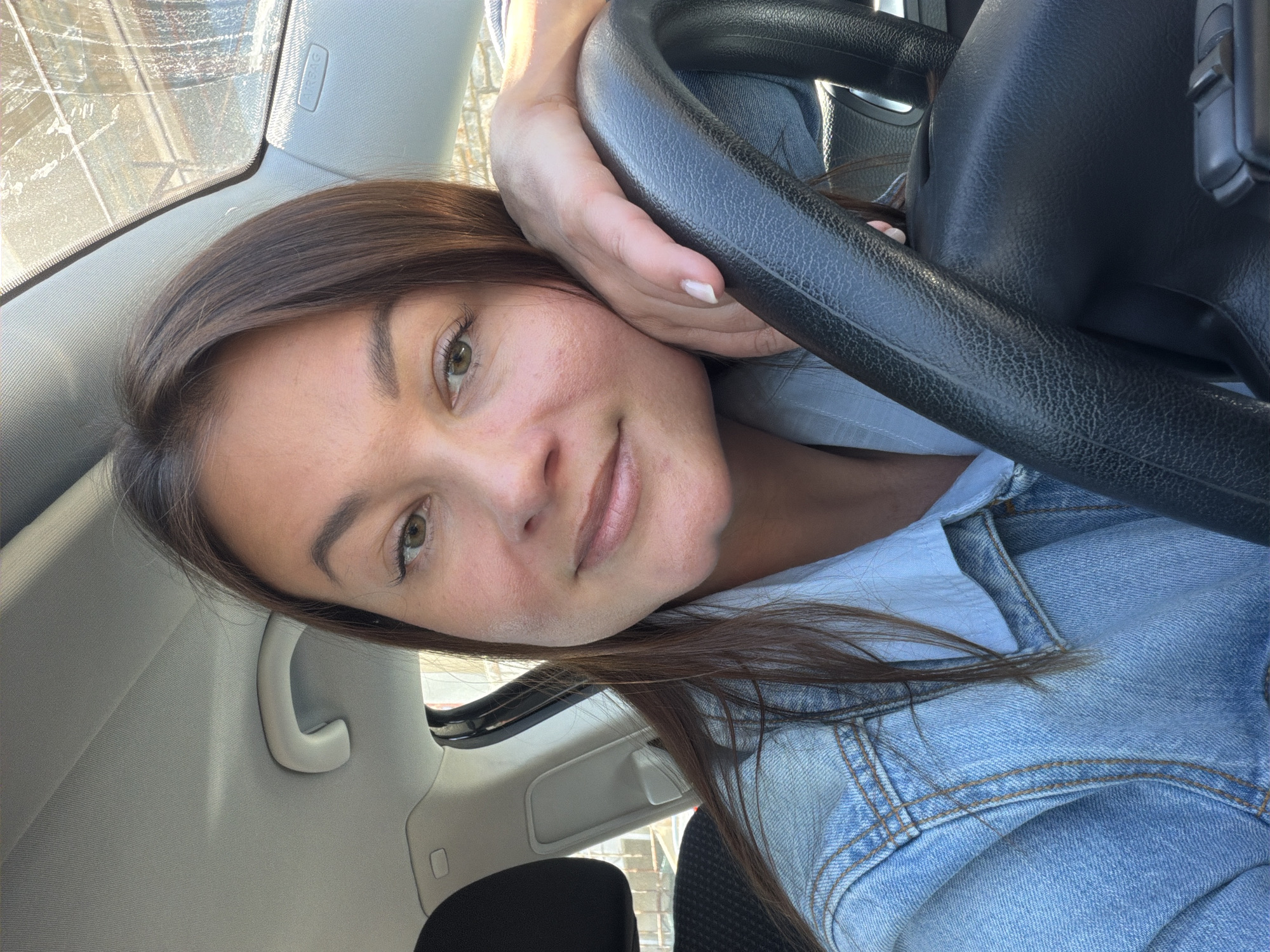 A woman with brown hair and light skin is pictured from a low angle inside a vehicle. Her left cheek rests on her left hand which is positioned on the steering wheel. She is wearing a light blue denim jacket over a light blue shirt. The interior of the car is light gray. Outside the window, there is a blurred view of what appears to be urban architecture, potentially in Budva, Montenegro.