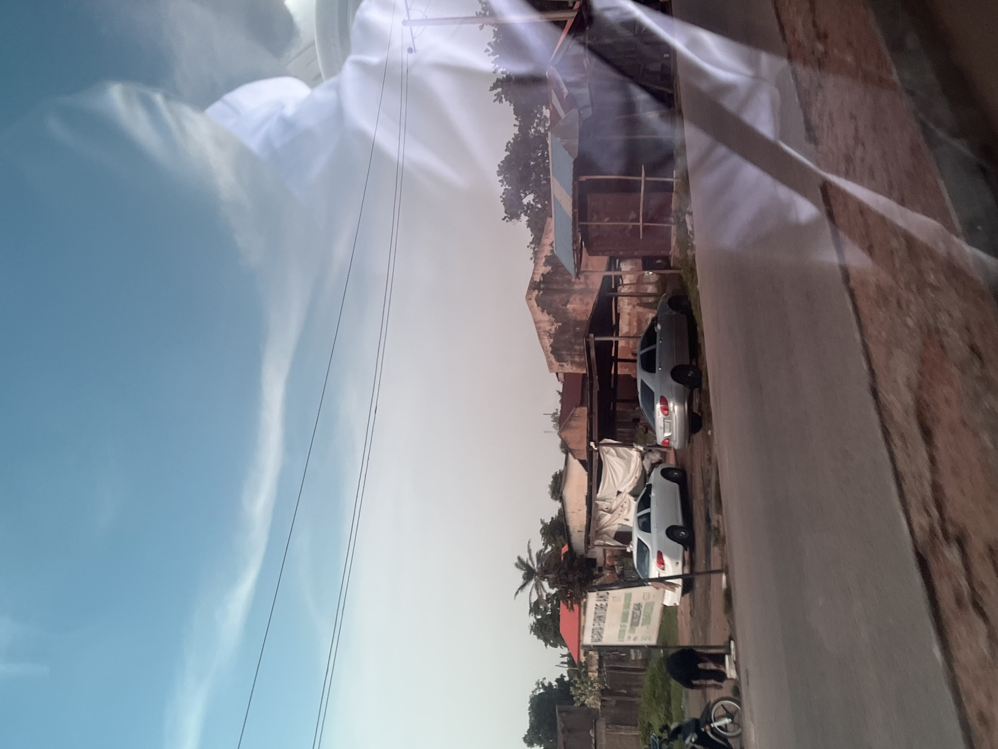 The image depicts a street scene in Jalingo, Nigeria, as seen through a reflective surface, likely a car window. The scene is oriented vertically, making it appear as if viewed from above, looking down. In the foreground, a blurred, bright white object, possibly part of a vehicle's interior or a reflection, dominates the left side. To the right, a gray asphalt road runs diagonally. Along the side of the road are several buildings with corrugated metal roofs and exposed brickwork.
Parked on the side of the road are two white cars. One building has a white tarp draped over it, suggesting ongoing construction or a temporary cover. In front of some of the buildings, there are signs. One sign, predominantly green and white, is partially visible and appears to have text in a language common in Nigeria. Trees and foliage are interspersed between the buildings, with a prominent palm tree visible towards the center-right of the frame.
In the lower right portion of the image, a person is partially visible, bent over near a motorcycle. The sky is a pale blue with wispy white clouds, indicating it might be late afternoon or early morning. The overall impression is of a somewhat dusty and warm environment, typical of a Nigerian town. The reflections on the viewing surface obscure some details and add a layer of distortion to the scene.