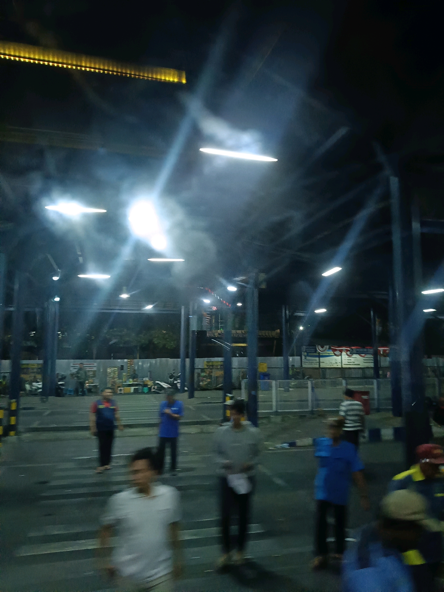 The image depicts an outdoor scene at night, likely a public gathering or transport hub, illuminated by overhead lights. In the foreground, several individuals are visible, some in motion and others standing. One person in a white shirt is in sharp focus, while others are blurred due to movement or distance. The background shows a covered structure supported by pillars, with various signs and what appear to be stalls or booths. A strip of yellow lights runs along the top of the structure. Several motorcycles are parked in the background. The overall atmosphere is busy and somewhat chaotic, suggested by the motion blur and the density of people and objects. The location is identified as Kedungrejo, Indonesia.