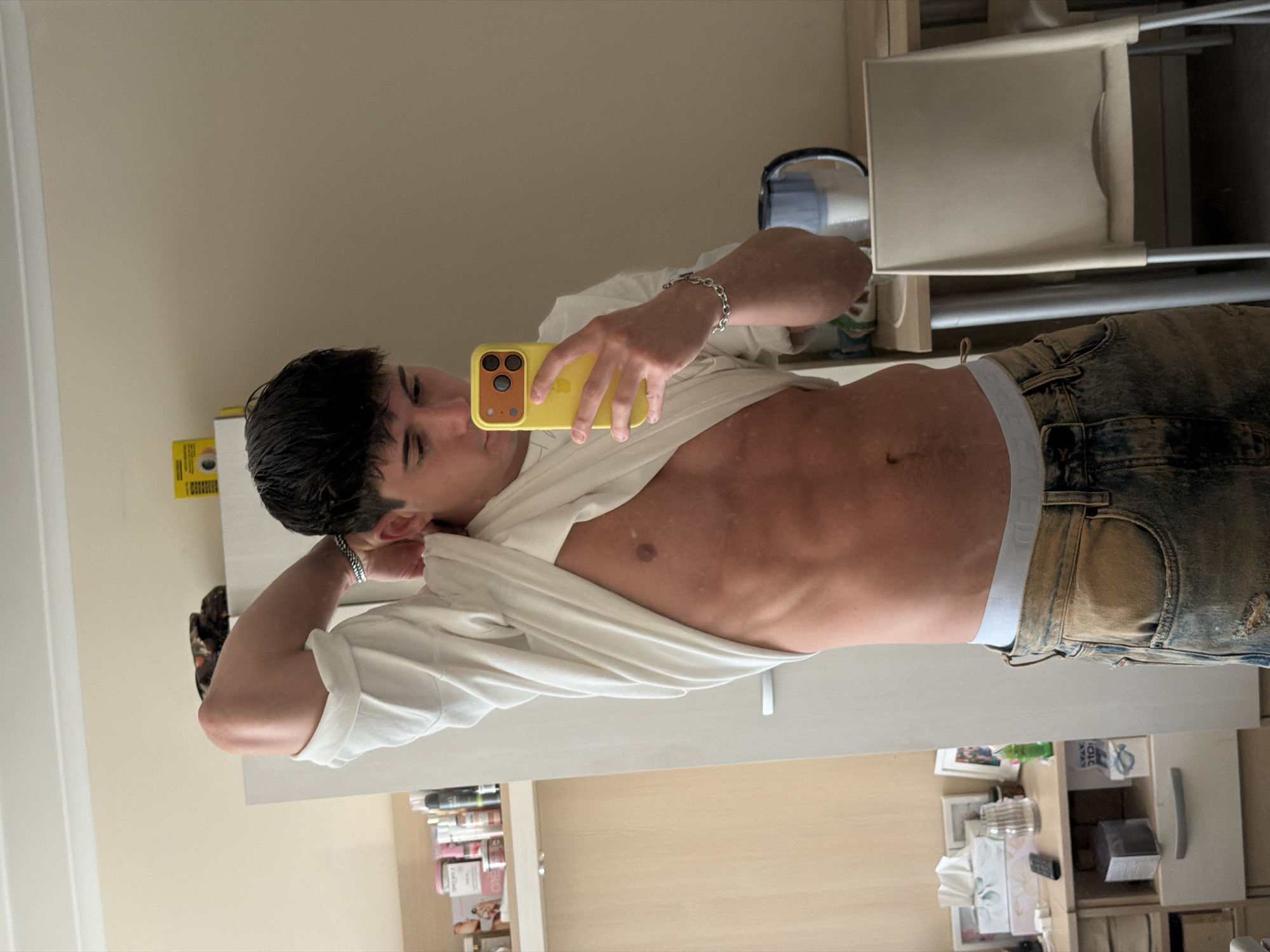 A slender, tanned man takes a selfie in a bathroom or bedroom. He holds a yellow-cased smartphone with three lenses in his right hand, obscuring much of his face. He wears a loose-fitting white shirt that is unbuttoned to reveal his abdomen and part of his chest. His hair is short and dark. The man also wears distressed jeans. The background features bathroom items on shelves and the bottom of a white chair. It is not possible to determine the location within Sofia, Bulgaria, from the image alone.