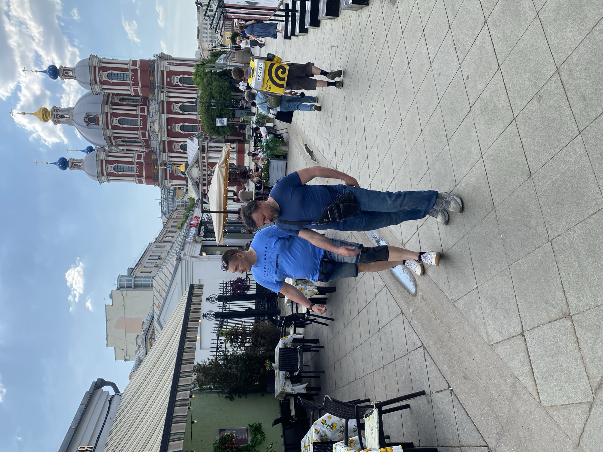 The image depicts a street scene in Moscow, Russia, with a red and white Orthodox church in the background. Two men are standing on a tiled sidewalk, one closer to the camera wearing a blue t-shirt and jean shorts, and the other further away wearing a blue t-shirt and jeans. They appear to be looking down. Behind them, other people are walking on the sidewalk. There's an outdoor seating area of a restaurant with tables and chairs on the left. A delivery person with a yellow backpack walks up the sidewalk. The sky is partly cloudy.