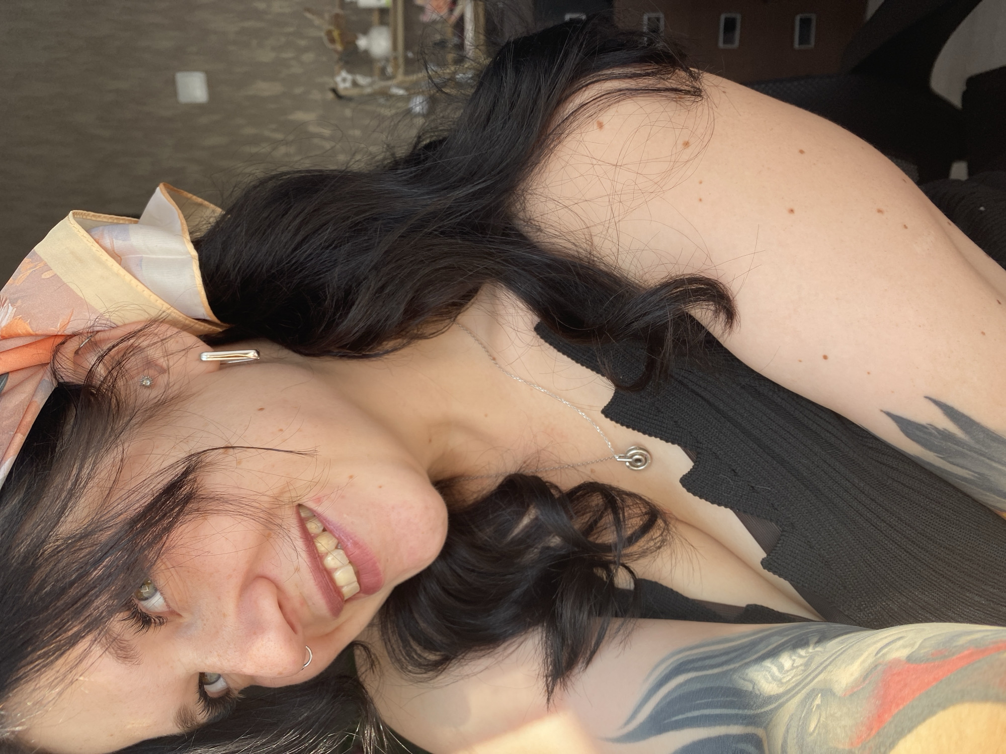 The image shows a close-up, upper-body portrait of a young woman with dark hair and fair skin, adorned with freckles. She is smiling, revealing slightly discolored teeth, and wearing a black, low-cut top. Her hair is styled in pigtails, and she has an orange and white patterned scarf tied around her head. She has a nose ring and wears silver earrings and a necklace with a pendant. Tattoos are visible on her left arm and shoulder. The background is out of focus, suggesting an indoor setting with muted colors and some shelving in the distance. The lighting indicates a sunny day, with a strong light source to her left. There is no discernible information in the image to suggest a location in the city of Chaikovsky, Russia.