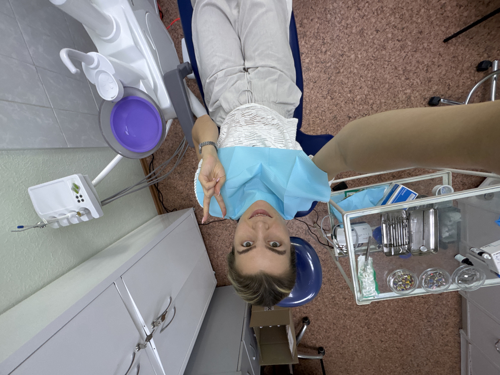 A woman, wearing a light-colored top and light-colored pants, is seated in a dental chair. She is wearing a light blue dental bib. Her left arm is raised, with her hand in a peace sign gesture. The woman's hair is blonde and pulled back. She is looking towards the camera with a slight smile. The scene is in a dental operatory. In the background, there is a dental unit with a purple bowl and dental instruments visible on a tray. A white cabinet is also present. The floor is cork-tiled. The image is taken from a high-angle perspective.