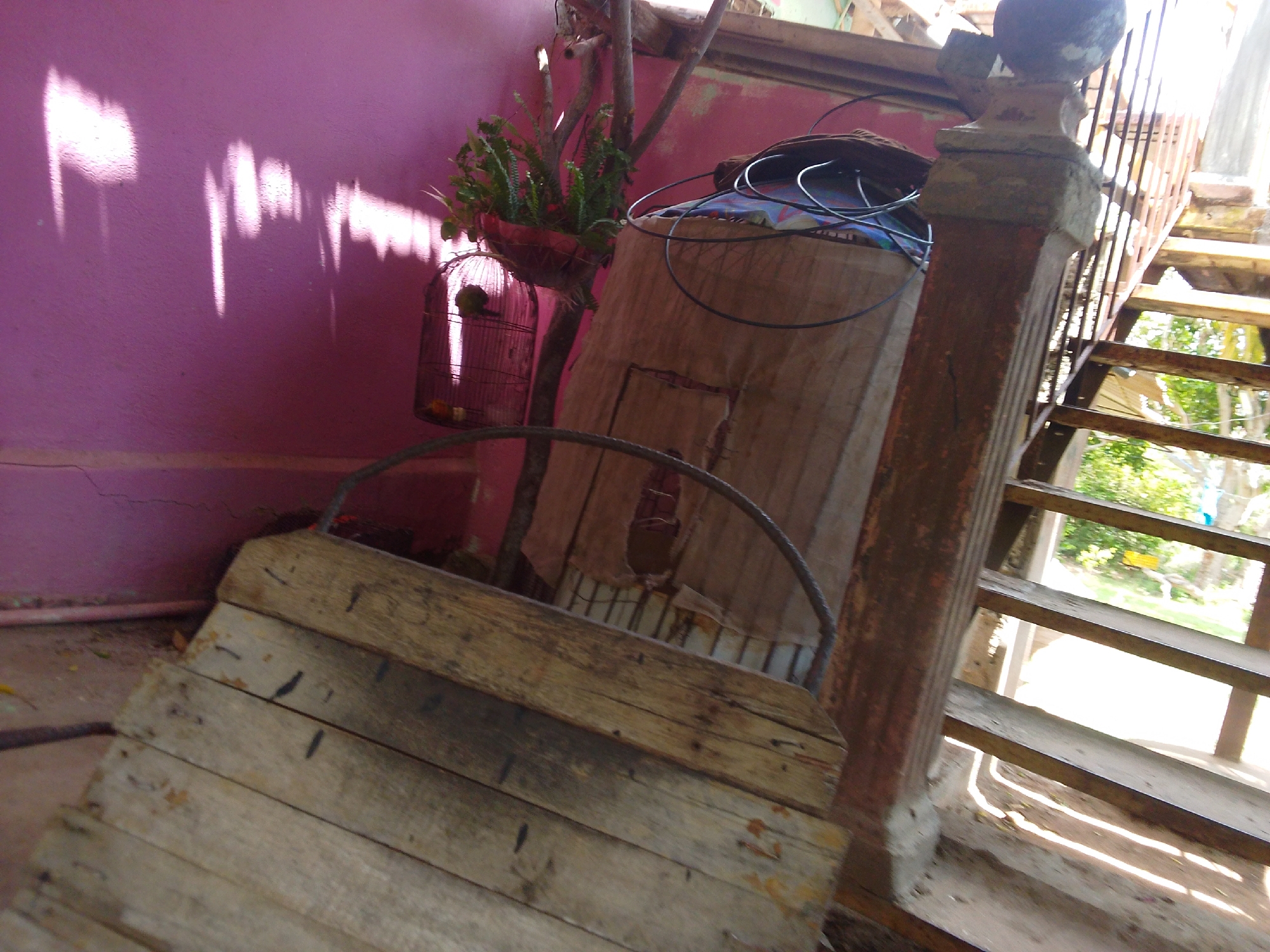 The image depicts an outdoor scene, likely in a residential area in Guayacán, Venezuela, characterized by rustic elements and natural light. In the foreground, a weathered wooden structure, possibly a ramp or a part of a makeshift staircase, is prominently featured. Its surface is made of several wooden planks, showing signs of age and use, with visible nails and dark markings.
To the left, a pink wall serves as a backdrop, with abstract patterns of light and shadow cast upon it, suggesting sunlight filtering through an unseen opening or structure. Adjacent to the wall, a birdcage hangs from a wooden post, containing at least one small bird and some food items. The cage is made of dark metal bars and has a round base. Above the cage, a potted plant with lush green foliage is attached to the same post.
Behind these elements, a tall, wooden structure, possibly a staircase railing, with vertical slats, is visible. A carved stone pillar, weathered and stained, stands next to the staircase. A dark, coiled cable rests on top of a large, fabric-covered object, which appears to be a bundle or a piece of furniture draped with burlap or a similar material. The overall impression is one of a lived-in, possibly slightly neglected, outdoor space in a warm climate. The lighting suggests it is daytime, with strong sunlight creating sharp shadows.