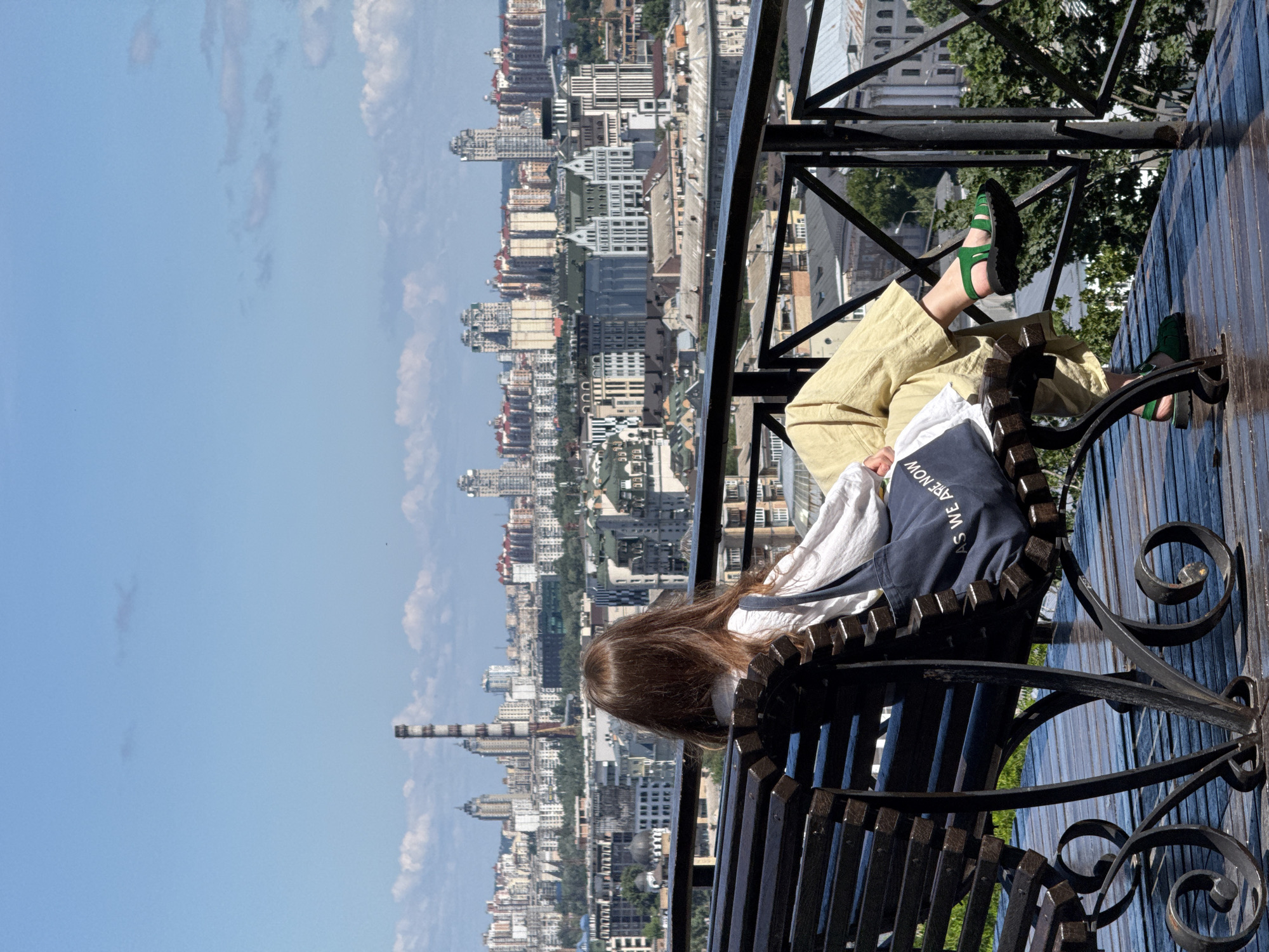A woman with long brown hair sits on a dark wooden bench, facing away from the camera, looking out at a panoramic view of Kyiv, Ukraine. She is wearing a white shirt and light yellow pants, and green sandals. A dark blue tote bag with white lettering that reads "AS WE ARE NOW" is slung over her shoulder and rests on the bench. The bench is made of dark wood and ornate metalwork. In the background, the cityscape of Kyiv stretches out under a clear blue sky with scattered white clouds. Prominent features in the skyline include a tall, red-and-white striped industrial chimney on the left, and numerous modern apartment buildings and other structures. A black metal railing runs along the edge of the elevated viewing platform, separating it from the city below. Green trees are visible in the lower right portion of the image. The overall scene captures a moment of quiet contemplation against the backdrop of a bustling urban environment.