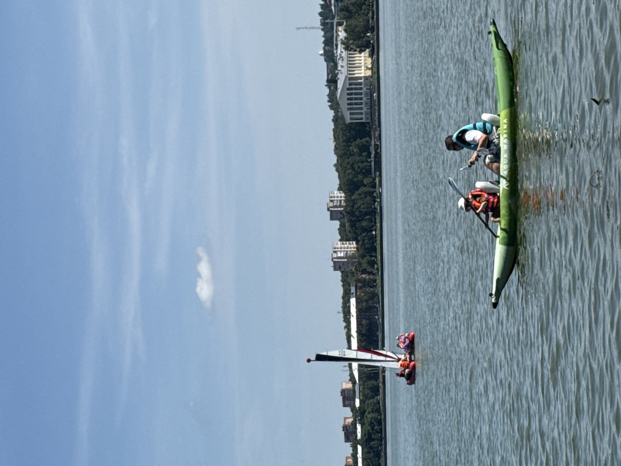 The image depicts a sunny day on a body of water, likely the Votkinskoye Reservoir, in Votkinsk, Russia. In the foreground, a green double kayak carries two individuals. The person in the back, a male wearing a blue and white life jacket, is paddling. The person in the front, a child wearing an orange life jacket and a white hat, is also holding a paddle. The kayak is labeled "AQUA MARINA."
Further in the distance, a small sailboat with red and white sails is navigating the water. It appears to be a catamaran-style boat with multiple people on board, all wearing life jackets, some in orange and pink.
The background reveals a treeline along the shore, beyond which are several buildings. One prominent building, with a classical facade featuring columns, is visible. Other buildings, including apartment complexes, are also scattered along the horizon. The sky is blue with a few wispy clouds. The overall scene is one of recreational activity on a calm body of water under clear skies.