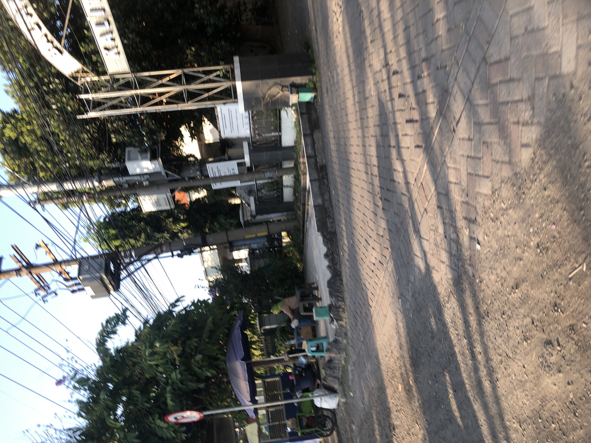 The image depicts a street scene in Kludan, Indonesia, likely during daylight. In the foreground, a paved walkway with a cobblestone-like pattern is visible, intersected by a dirt or gravel path. Sunlight casts long shadows across the ground, suggesting either early morning or late afternoon.
On the left side of the image, a small stall with a blue umbrella is set up. A cart with wheels, possibly a food cart, is parked nearby. Two individuals are present at the stall; one appears to be sitting on a stool, while the other is hunched over, possibly working. A green plastic stool and a blue container are also at the stall. A red and white circular sign with a diagonal line through it, likely indicating a prohibition, is mounted on a pole.
In the background, a utility pole with numerous wires and a transformer is prominent. Further back, an archway with metal supports spans across the street. A sign with white lettering, partially obscured and blurry, is visible on the archway, reading "KAL...". Trees with dense green foliage line the background, and a building with a white facade can be seen behind the archway. A dark-colored trash receptacle is situated near the base of the archway on the right. The overall impression is a moderately busy street environment with commercial activity and infrastructure.