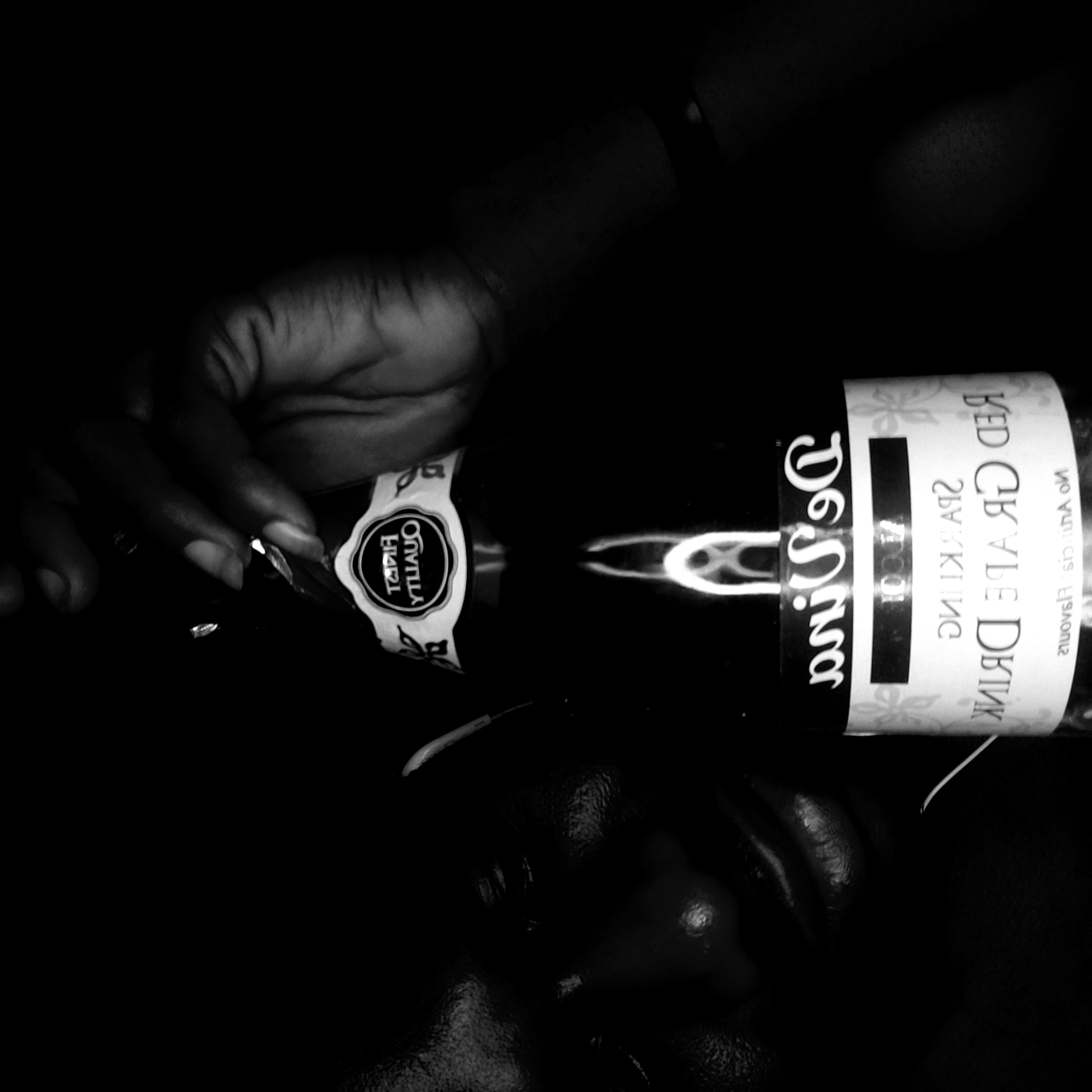 A black and white, horizontally oriented image depicts a partially visible person's face at the bottom, with a hand holding a bottle of "DeSina Red Grape Drink" diagonally across the frame. The background is dark and indistinct. The label on the bottle is partially visible, displaying text in reverse. The face and hand are in dark tones, creating contrast with the bottle's label. The presence of "DeSina" brand suggests potential origin or association within Nigeria, but there is no evidence to confirm the image was taken in Narayi.