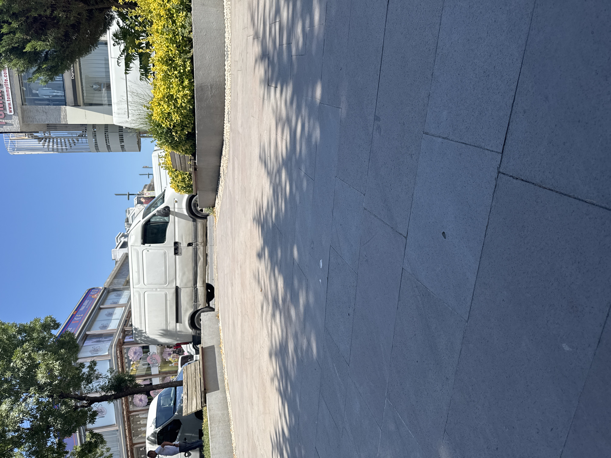 A daytime, eye-level shot depicts a street scene in Gungoren, Turkey, characterized by modern architecture and a clear blue sky. In the mid-ground, a white van is parked. To its left, a white passenger van is also parked, and a man in a white t-shirt and blue jeans stands beside it, facing away from the camera. In the background, the facade of a multi-story building with large windows and a spiral staircase is visible. Next to it, another building features shops with mannequins displayed in the windows. A lush green bush lines the edge of a raised planter in the mid-ground. The foreground consists of a paved walkway with large, grey stone tiles, upon which dappled shadows from trees are cast. The overall impression is one of an urban commercial area on a bright, sunny day.