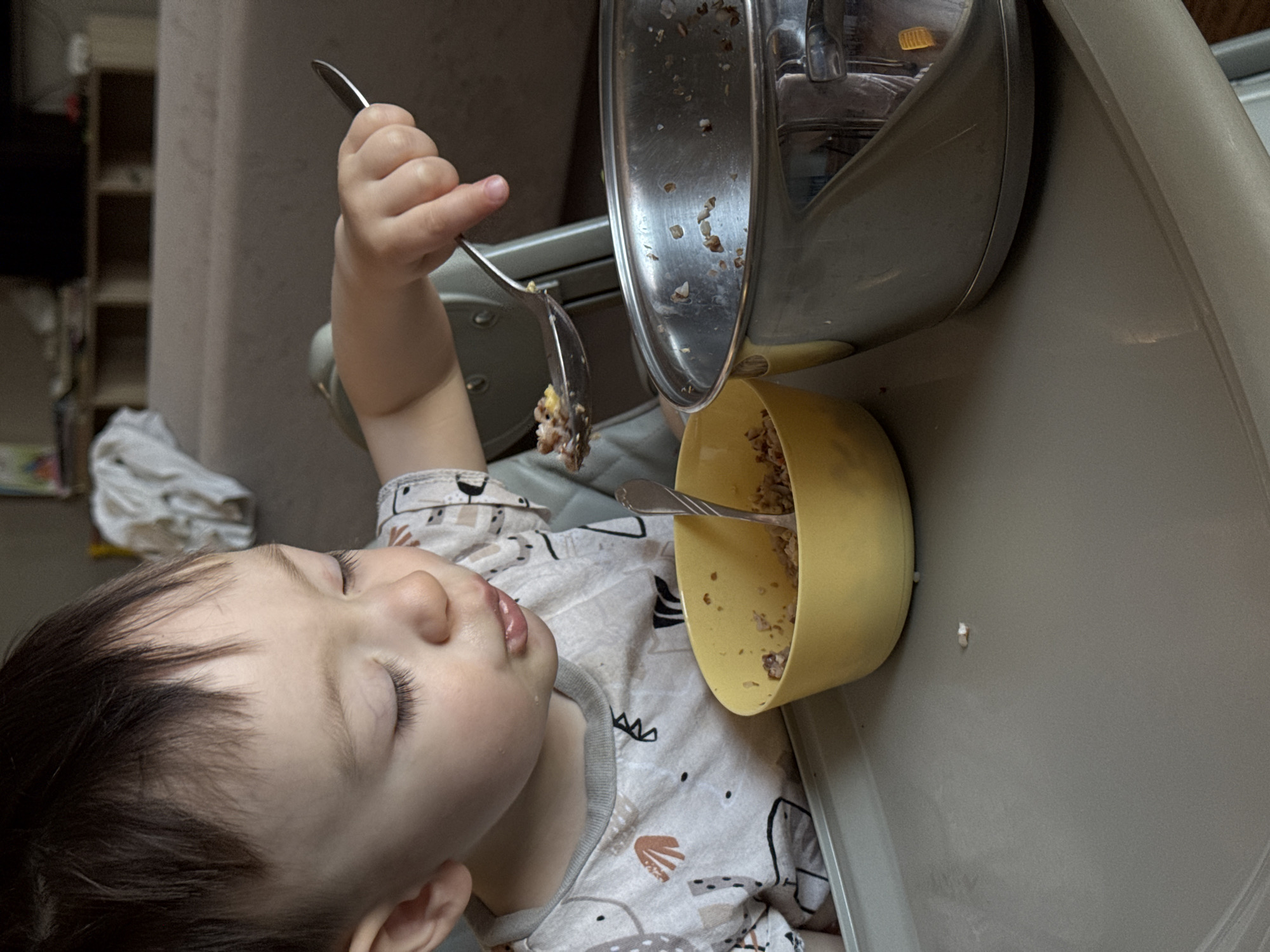 A baby is feeding itself from a yellow bowl on a high chair with a metal tray. The baby is holding a silver spoon with food on it.