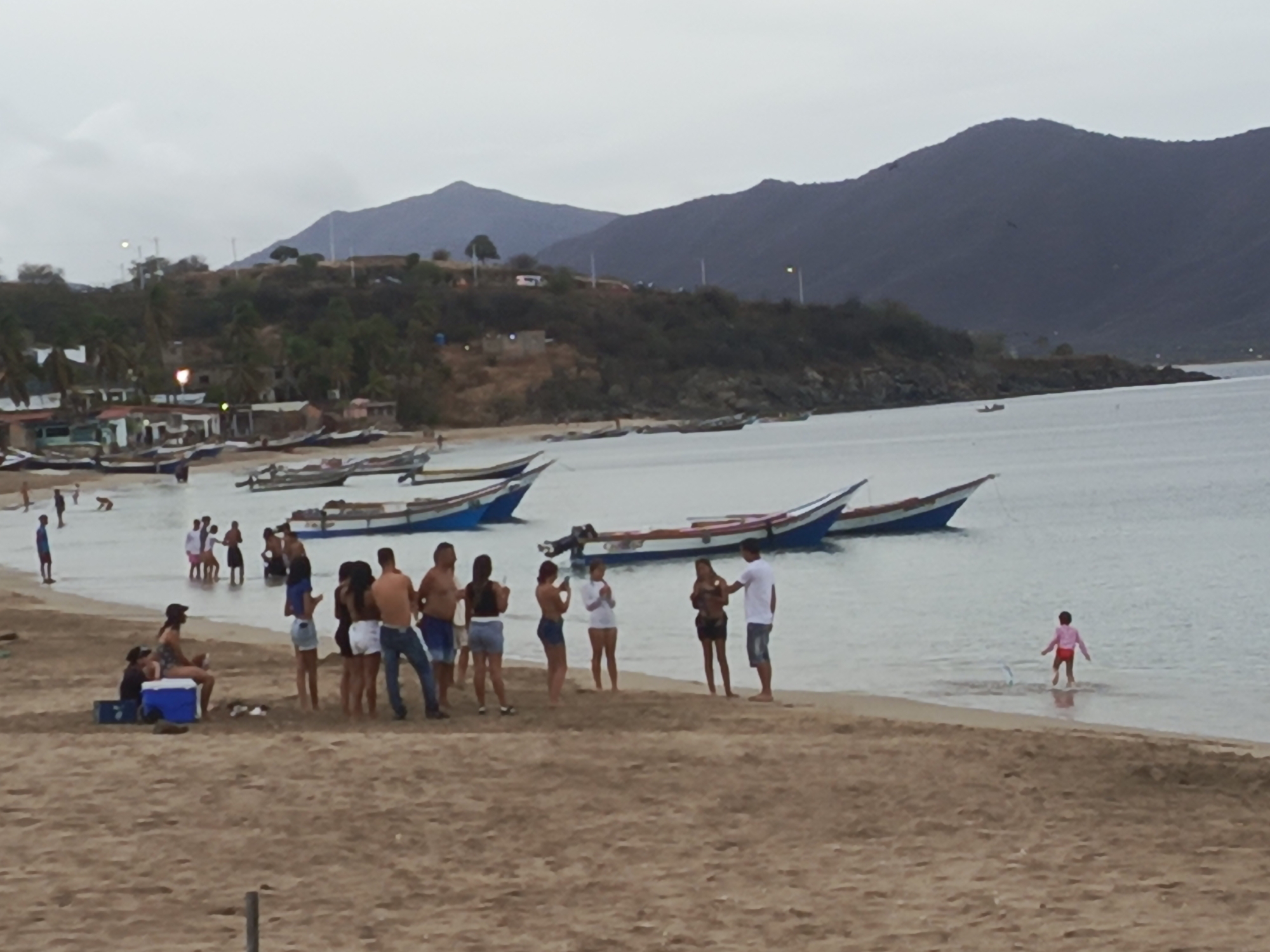 The image shows a beach scene in Juan Griego, Venezuela. In the foreground, there is a sandy beach with several people gathered. Some individuals are standing and talking, while others are sitting. A blue and white cooler is visible near a seated person. Further back, a group of people are in the shallow water. Several boats are moored in the bay, and in the background, there are hills and mountains. The sky is overcast.