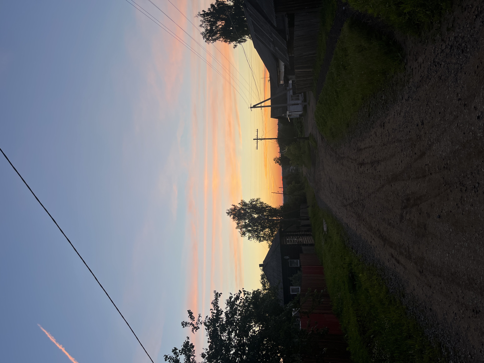 This image depicts a rural road in Borodulino, Russia, at dusk. The sky is painted with hues of pink and orange, with a single contrail visible in the upper left quadrant. The road is unpaved and dusty, with tire tracks suggesting recent vehicle passage. On either side of the road, traditional wooden houses with dark roofs and fences are visible, some with satellite dishes. Trees and overgrown grass line the road and property edges, indicating a natural, somewhat unkempt environment. Power lines stretch across the sky, supported by wooden poles, adding to the rural atmosphere. The overall scene is serene, capturing the quiet beauty of a Russian village at the end of the day.