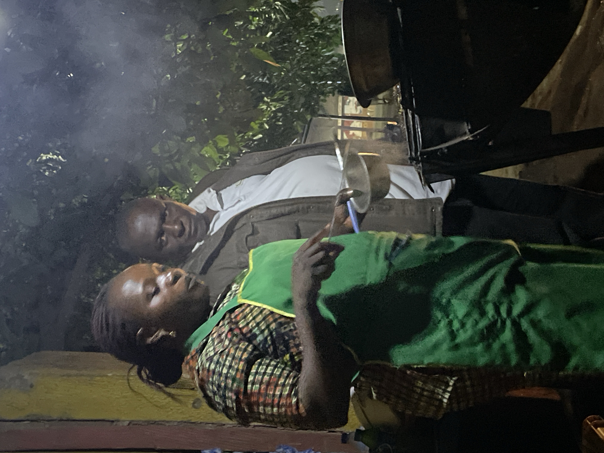 Two individuals, a man and a woman, are positioned outdoors at night with smoke visible in the scene. The woman, wearing a green apron over a patterned shirt, is seated and pointing her index finger upwards. Beside her, a man in a white shirt and brown jacket looks towards the woman. Above them, there is a large cooking pot mounted on a stand. The location is Jos, Nigeria, based on available context.