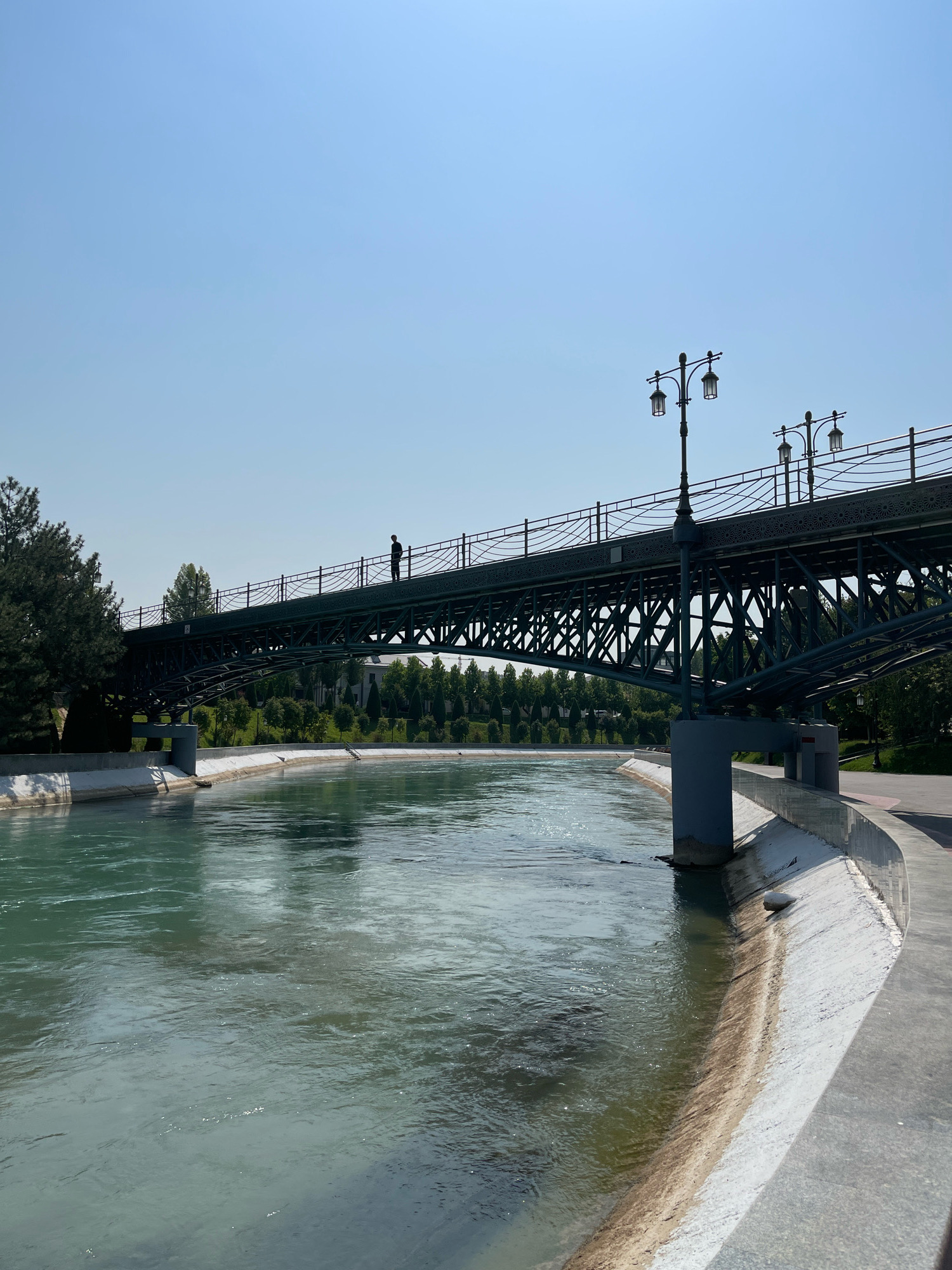 A man stands silhouetted on an arched pedestrian bridge over a wide, greenish-blue canal in Tashkent, Uzbekistan. The bridge, made of dark metal with intricate filigree work along its railings and supporting structure, is illuminated by several ornate street lamps. Trees line the far bank of the canal, with some buildings visible behind them. The near bank features a concrete walkway and a sloping, concrete embankment leading into the water. The sky is clear and bright blue.
