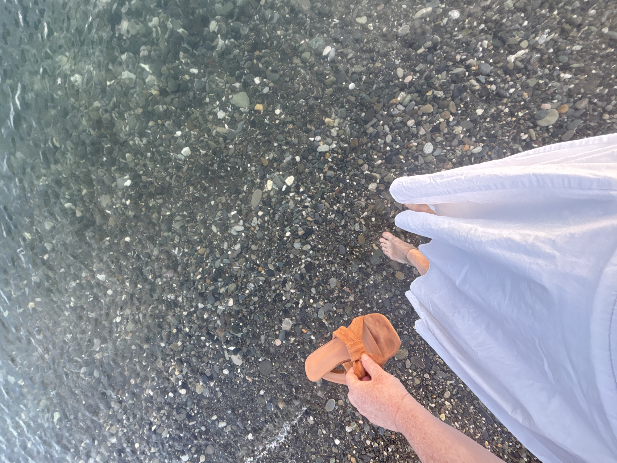 In the city of Sochi, Russia, a person stands at the edge of the Black Sea. Their legs are partially submerged in the clear, shallow water, revealing a bottom covered in smooth, dark pebbles. A white skirt drapes over their legs. In their right hand, they hold a single orange sandal. The scene is captured from a high-angle perspective, looking down onto the water and the shoreline.