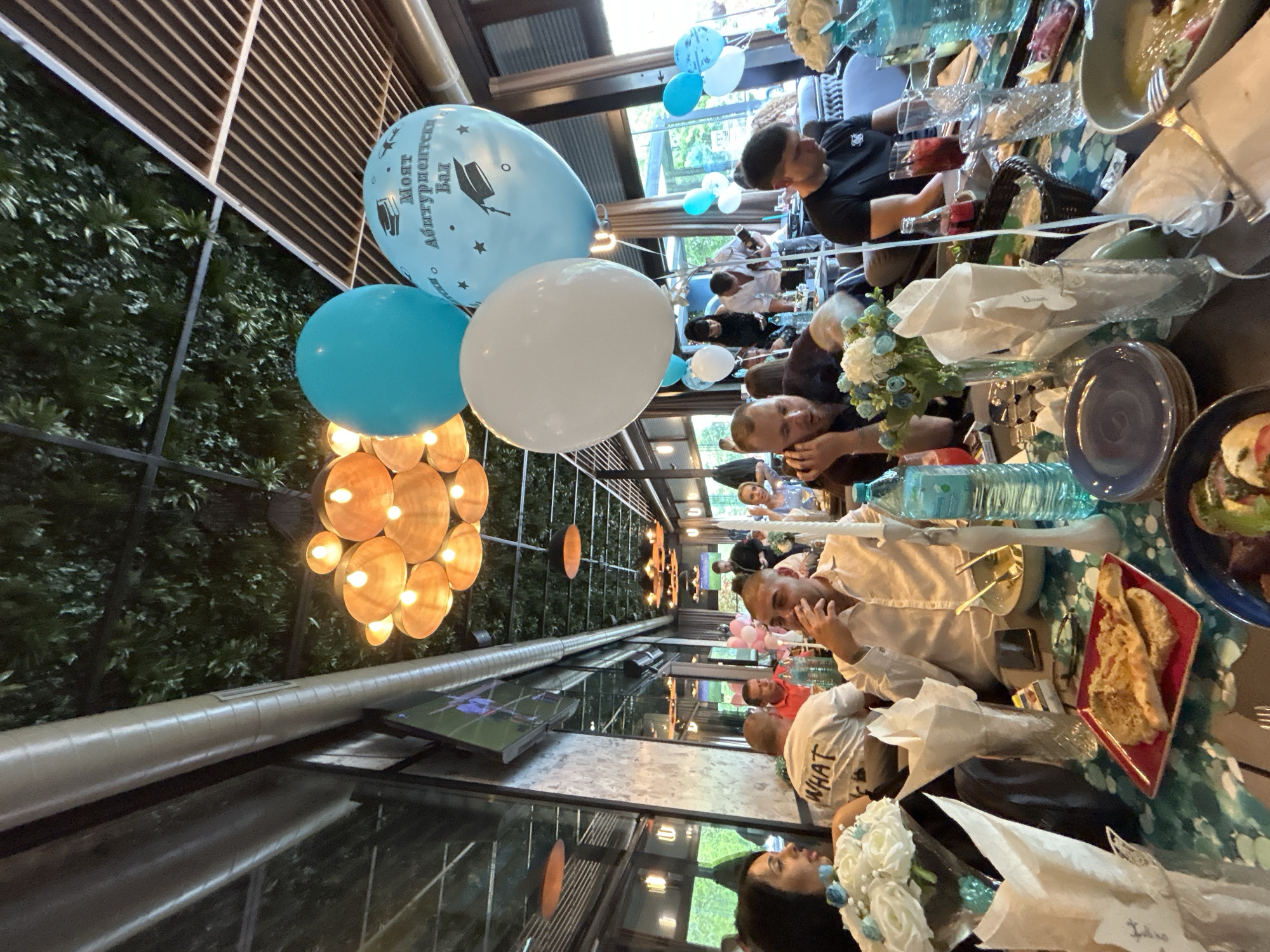 The image depicts an indoor event, possibly a celebration, at a restaurant or event space, judging by the tables and decorations. There are people seated at the tables, engaged in conversation and enjoying food and drinks. Balloons with graduation motifs are visible, suggesting a graduation party. The location appears to be a modern, enclosed patio or garden-style setting with greenery and decorative lighting. There is a TV screen visible in the background. The presence of Bulgarian text on one of the balloons suggests this is likely taking place in Bulgaria. The phrase on the balloon, "Моят Абитуриентски Бал," translates to "My Graduation Ball". Based on this information and the style of architecture and décor, it is highly probable that this event is taking place in Burgas, Bulgaria.