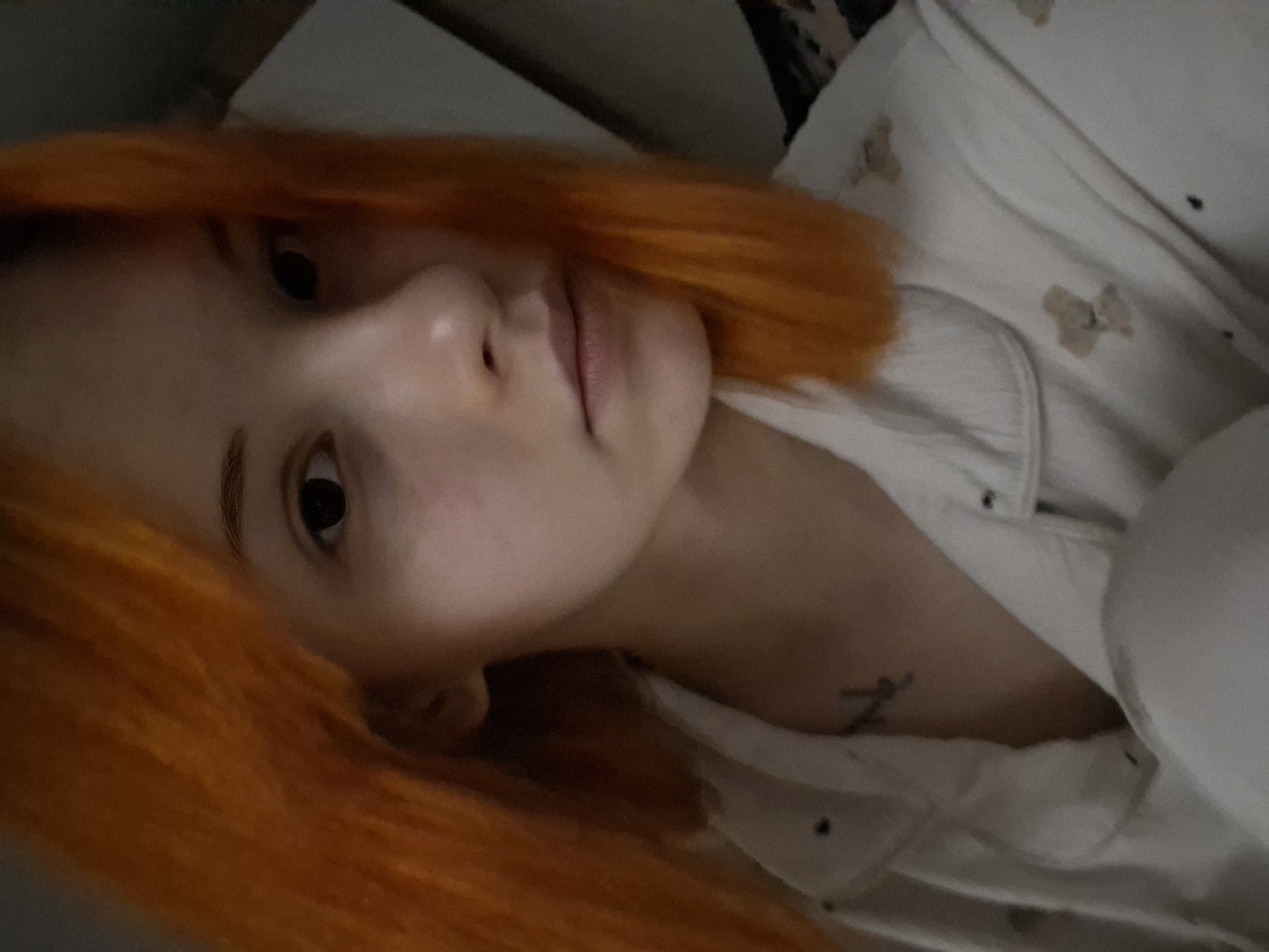 A close-up, portrait-style image features a person with striking, short orange hair, lying down and looking directly at the camera. Their head is tilted, suggesting they are resting on their side or back. The person has fair skin, dark eyes, and a neutral, relaxed expression with closed lips. They are wearing a white collared shirt or pajamas adorned with a repetitive pattern of small, light brown, teddy bear-like figures. A small, dark, stylized tattoo, possibly a letter or symbol, is visible on their right shoulder or upper chest, partially covered by the clothing. The lighting is dim and soft, indicative of an indoor setting, possibly a bedroom, without strong direct light. The background is mostly out of focus, showing soft white bedding and darker indistinct areas. The overall mood is calm and personal. The location context provided for this media is Chaikovsky, Russia.