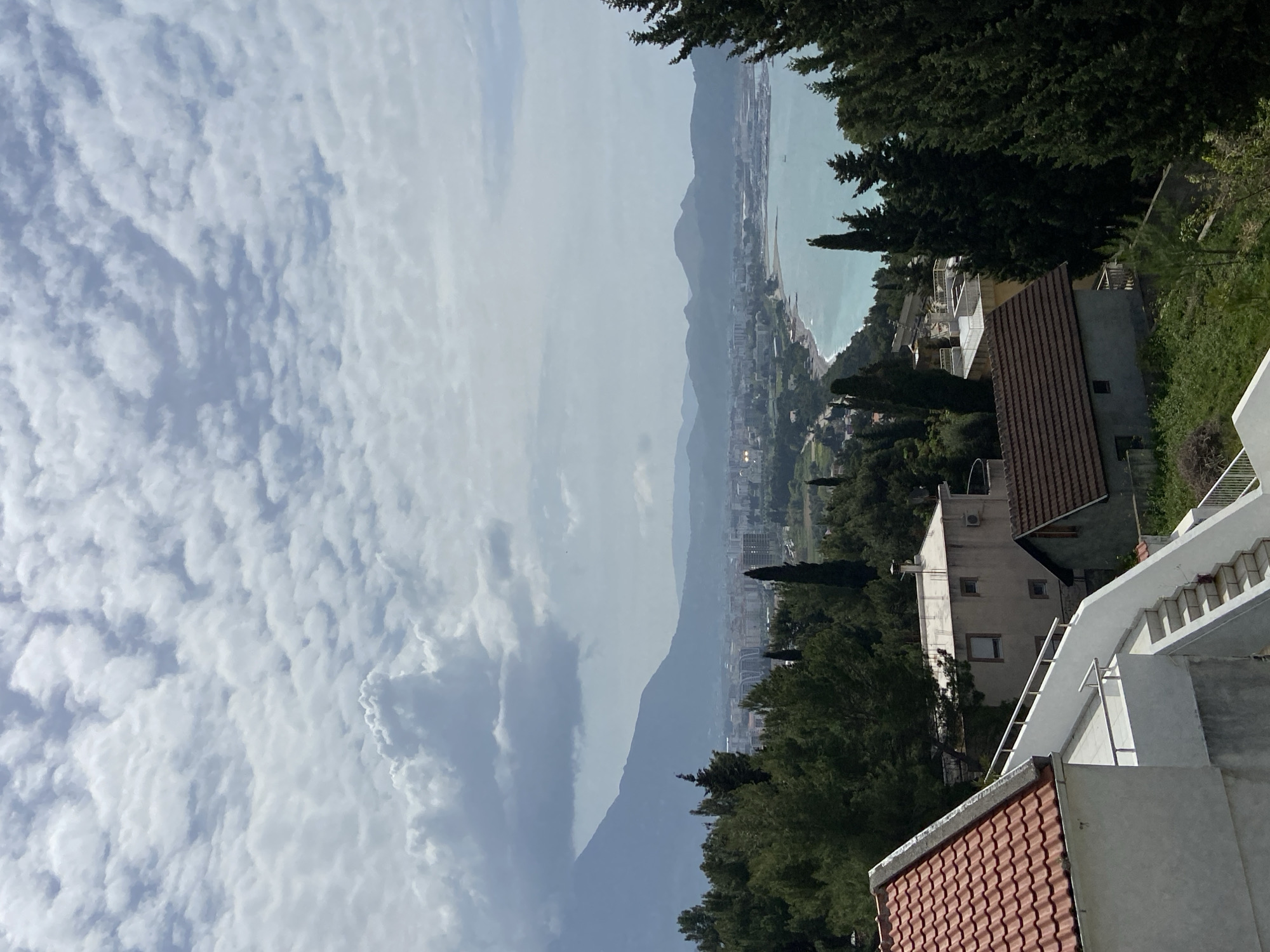 The image shows a coastal view from an elevated position in Šušanj, Montenegro. The sky is filled with a complex pattern of cumulus clouds. The coastline stretches into the distance, bordered by the Adriatic Sea. A line of buildings and urban infrastructure is visible along the coast. The foreground features residential buildings with terracotta tiled roofs and white concrete structures. Trees are scattered throughout the scene.