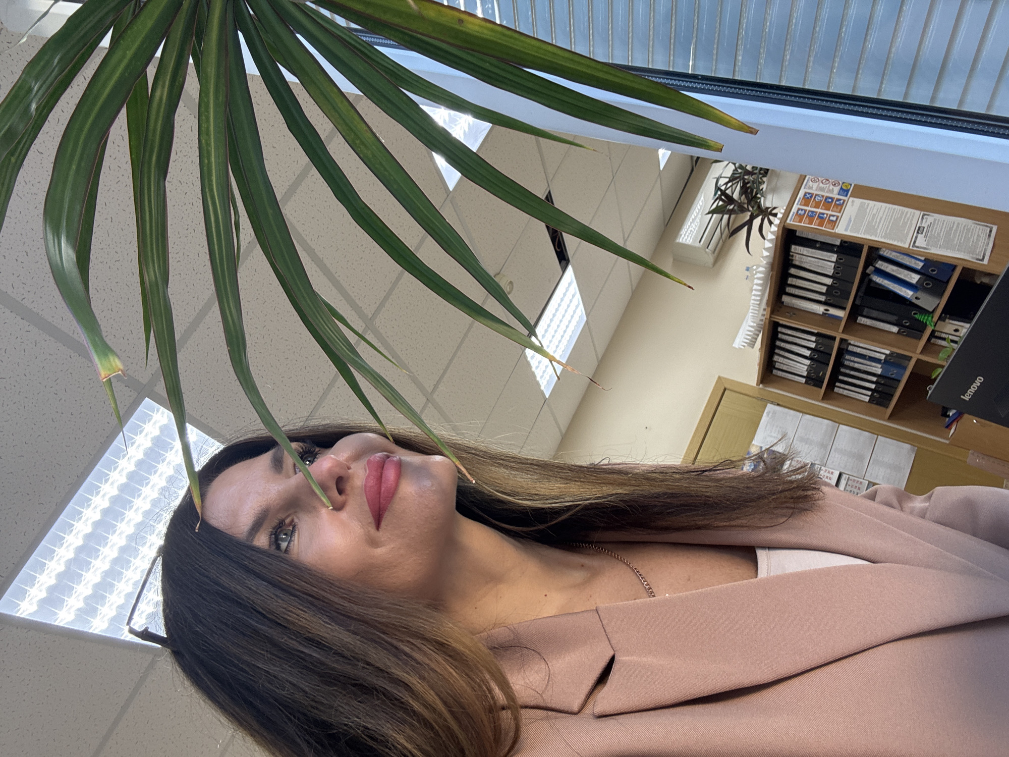 A medium shot, waist-up, captures a woman in an office setting. She is positioned in the foreground, facing left, with her head turned to look upwards and to the left. She has long, dark brown hair, parted and falling over her shoulders and down her back. Her makeup includes lipstick in a bright pink shade and visible eyelashes. She is wearing a light pink blazer over a white top, and a thin rose gold necklace is visible around her neck.
In the background, a shelving unit is visible, filled with binders and folders. To the right of the woman, a large plant with long, green leaves partially obscures the view of the background. The environment appears to be an office, with fluorescent lighting panels visible on the ceiling and what appears to be a window with blinds on the right side of the frame. A white air conditioning unit is mounted high on the wall in the background. The overall impression is that of a personal photograph taken within an office space. The image does not provide any specific information to confirm a location within the city of Chaikovsky, Russia.