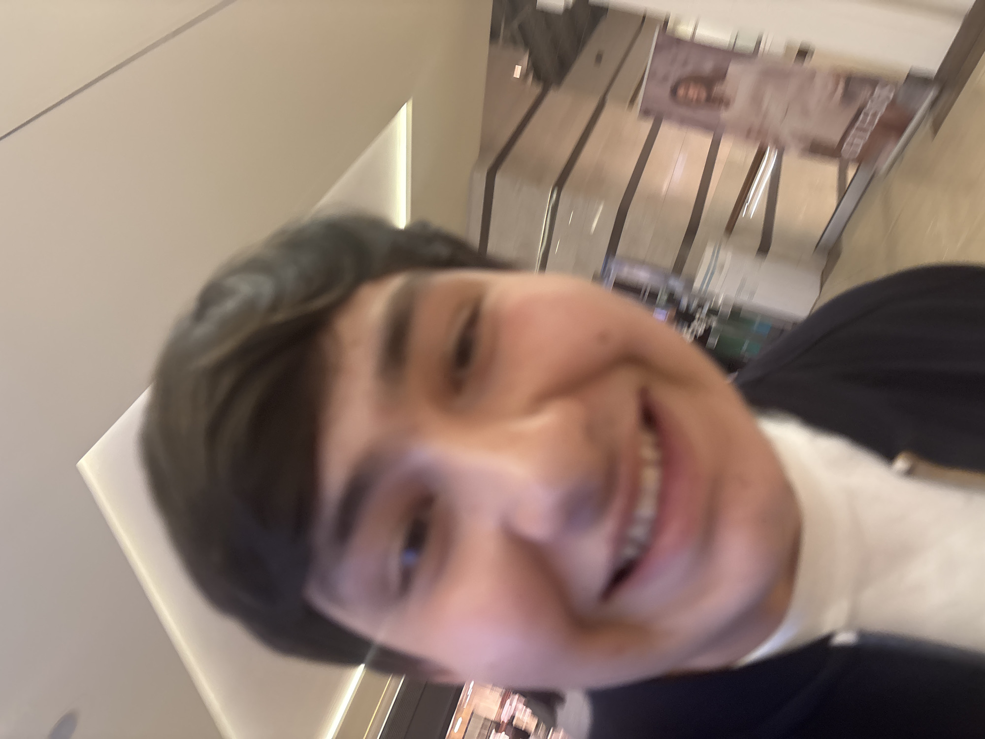 A close-up, angled selfie of a young man with short, dark hair wearing a white shirt and dark jacket. He is smiling. The background appears to be an indoor space with a store display and a hallway. While the location in the city of Yerevan, Armenia, is unidentifiable from the information provided in the image, the subject is in a building.