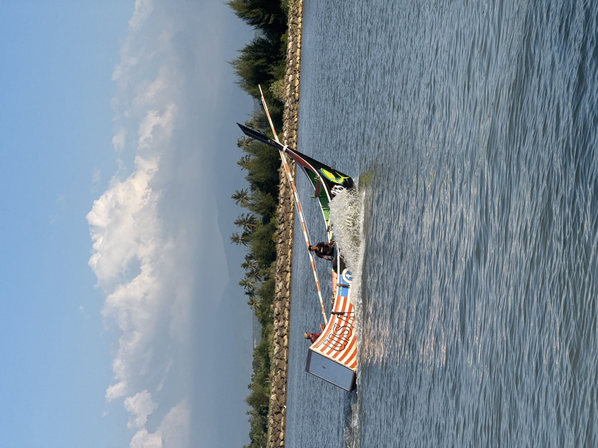 Two individuals are aboard a narrow, colorful fishing boat as it speeds through the water, creating a significant wake. The boat, decorated with orange and white stripes and featuring an image of a fish near its prow, is tilted to its port side. The person in the back, wearing a black shirt, is operating the boat. The person in the front, wearing a straw hat and a red shirt, is seated. The boat is in a body of water that appears to be a river or inlet, with a breakwater made of rocks and a line of palm trees and other foliage along the distant shore. In the background, hazy mountains are visible under a bright blue sky with scattered white clouds. The scene is set in Meunasah Jurong, Indonesia.