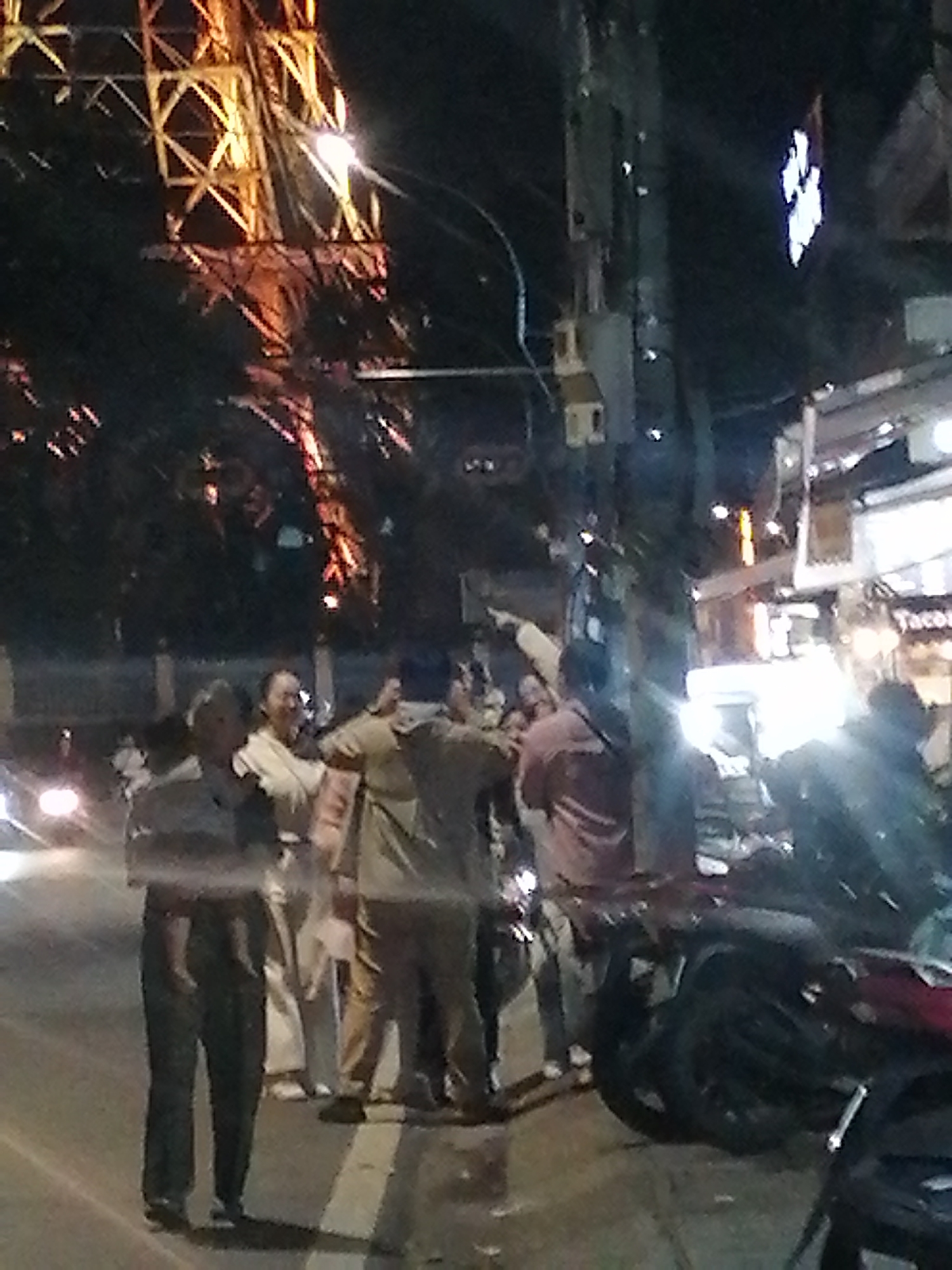 The image captures a nighttime scene on a street in Da Lat, Vietnam, with a prominent, illuminated structure resembling the Eiffel Tower in the background. In the foreground, several individuals are gathered near a utility pole. One person, wearing dark pants and a dark top, stands on the left, partially obscured by the pole. To their right, a person in a light-colored garment smiles. Behind them, two other individuals are visible, one in a tan outfit, and another in a pinkish shirt. Several motorcycles are parked on the right side of the street, and a sign with "Tacos" is visible above a building on the far right. The street appears to be paved, with a white line marking the edge of the road. The overall atmosphere suggests a public gathering or a busy street corner at night.
