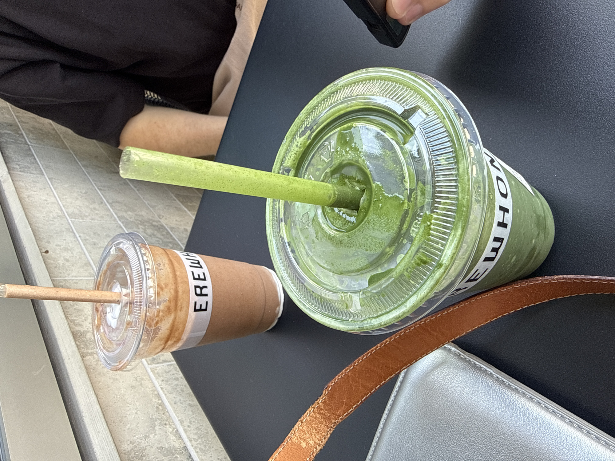 The image shows two Erewhon beverages on a black table. The drink on the left is in a brown paper cup with a clear plastic lid, and the word "EREWHON" is printed vertically on a white label around the cup. A light brown straw extends from the lid. The drink on the right is in a clear plastic cup with a clear plastic lid, and the word "EREWHON" is printed vertically on a white label around the cup. A green straw extends from the lid. The drink appears to be a green smoothie or juice. A brown leather purse strap and silver purse are visible in the lower right corner of the image. A person in dark clothing is partially visible in the upper left corner. The photo was taken outside, possibly on a patio, in West Hollywood, California, United States of America.