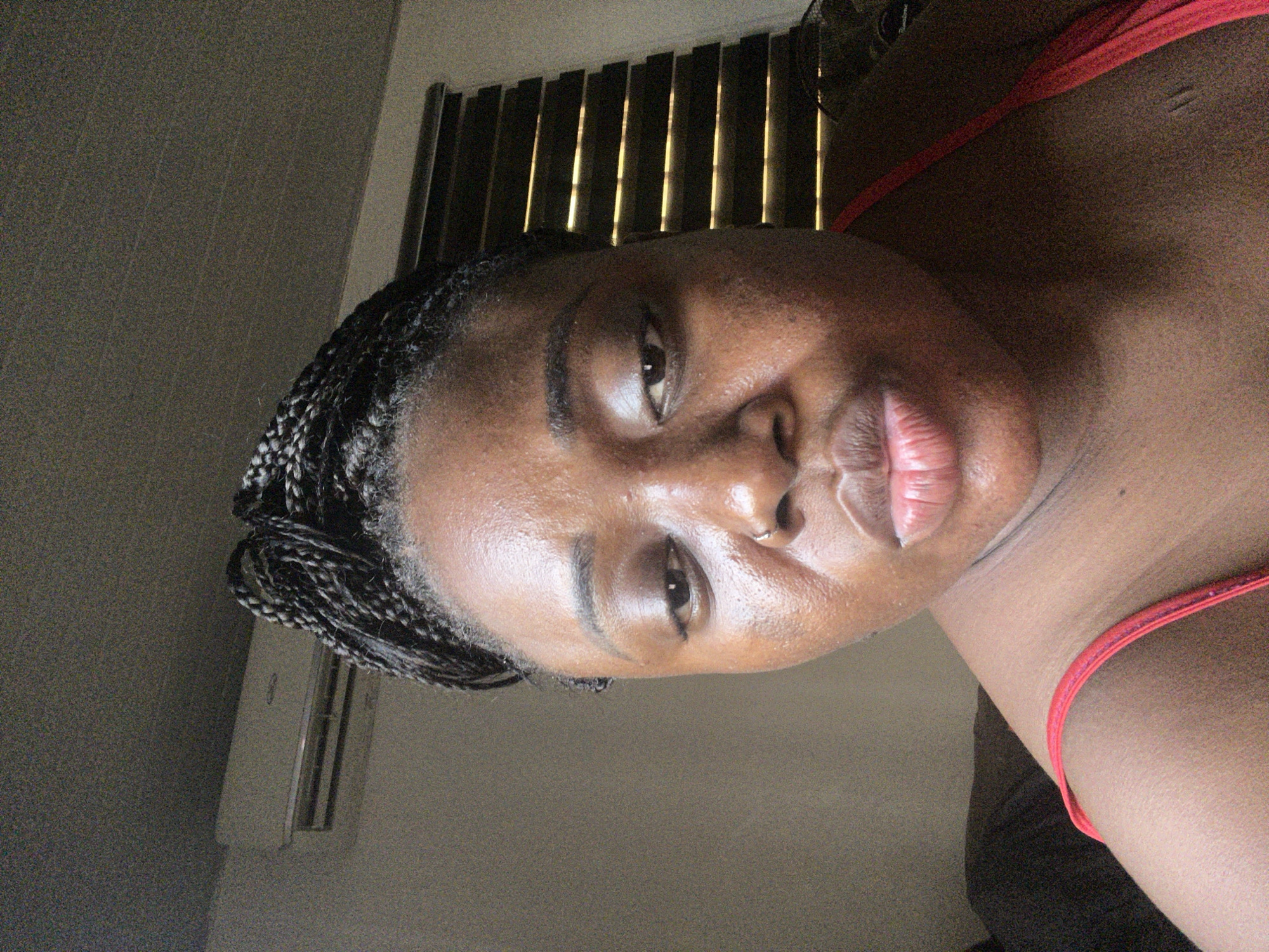 The image shows a close-up of a Black woman's face. She has dark hair styled in braids. She is wearing a red tank top. Behind her are closed window blinds with sunlight filtering through. An air conditioning unit is visible on the wall. It's impossible to ascertain the location within Abuja, Nigeria, from the provided image. There are no further details about actions, interactions, or other subjects.