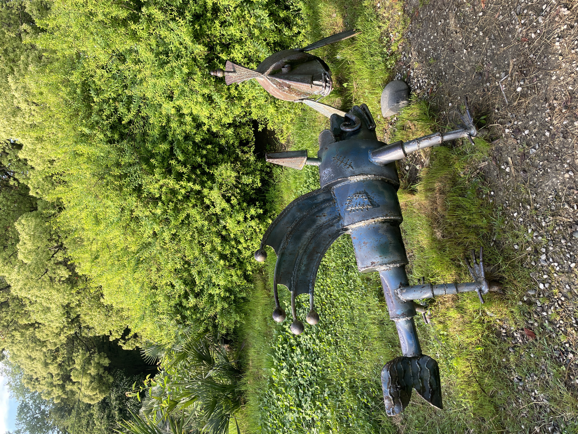 A metal sculpture stands on a grassy area next to a gravel path, with dense green foliage in the background. The sculpture is constructed from various metal pieces, including pipes, plates, and spheres. The metal has a weathered, blue-grey finish. The sculpture is possibly located in Sochi, Russia.