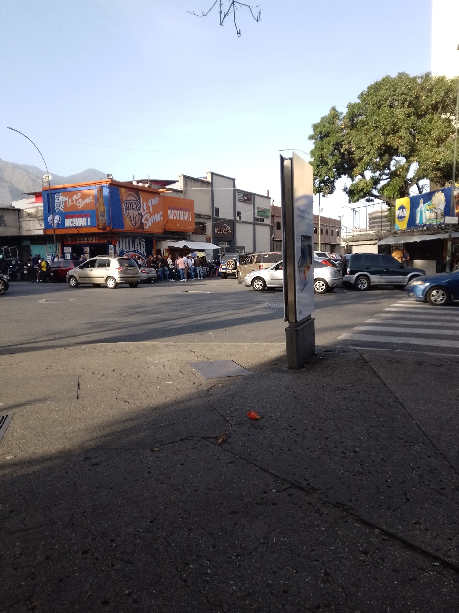The image depicts an urban street scene in Caracas, Venezuela. In the foreground, a sidewalk with cracked pavement leads to a pedestrian crosswalk. A metal advertisement pillar stands on the corner. Cars of various makes and models, including a silver hatchback, a dark SUV, and a blue sedan, are visible in the intersection. A crowd of people gathers near a building adorned with large "POLAR" and "NICOMARI I" signs, suggesting a commercial establishment or restaurant. In the background, mountains are visible beneath a clear sky. The scene indicates a moderately busy commercial area in Caracas.