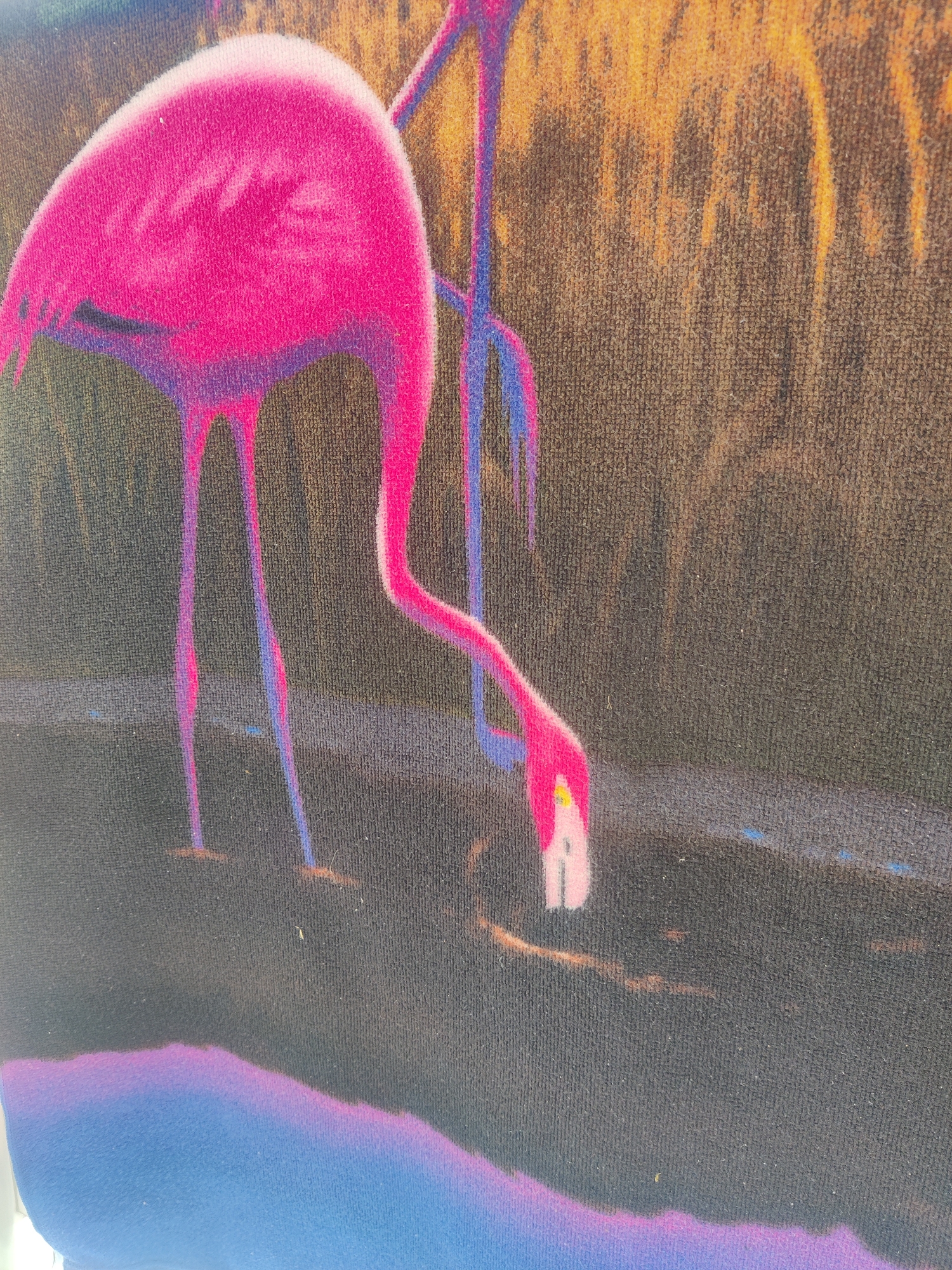 The image shows a close-up of a vibrant, abstract artwork featuring two flamingos. The flamingo on the left is predominantly pink, with its body taking up a significant portion of the frame. Its long, thin legs are visible, extending downwards into what appears to be water. The flamingo on the right is partially visible, with its legs also depicted. The background is a mix of warm, fiery oranges and yellows, suggesting a sunset or warm lighting, contrasted with darker, muted tones. The fabric texture is evident, indicating the artwork might be printed on a textile material. The location within Katerini Municipality, Greece, is not discernible from the image itself; this detail would require external information.