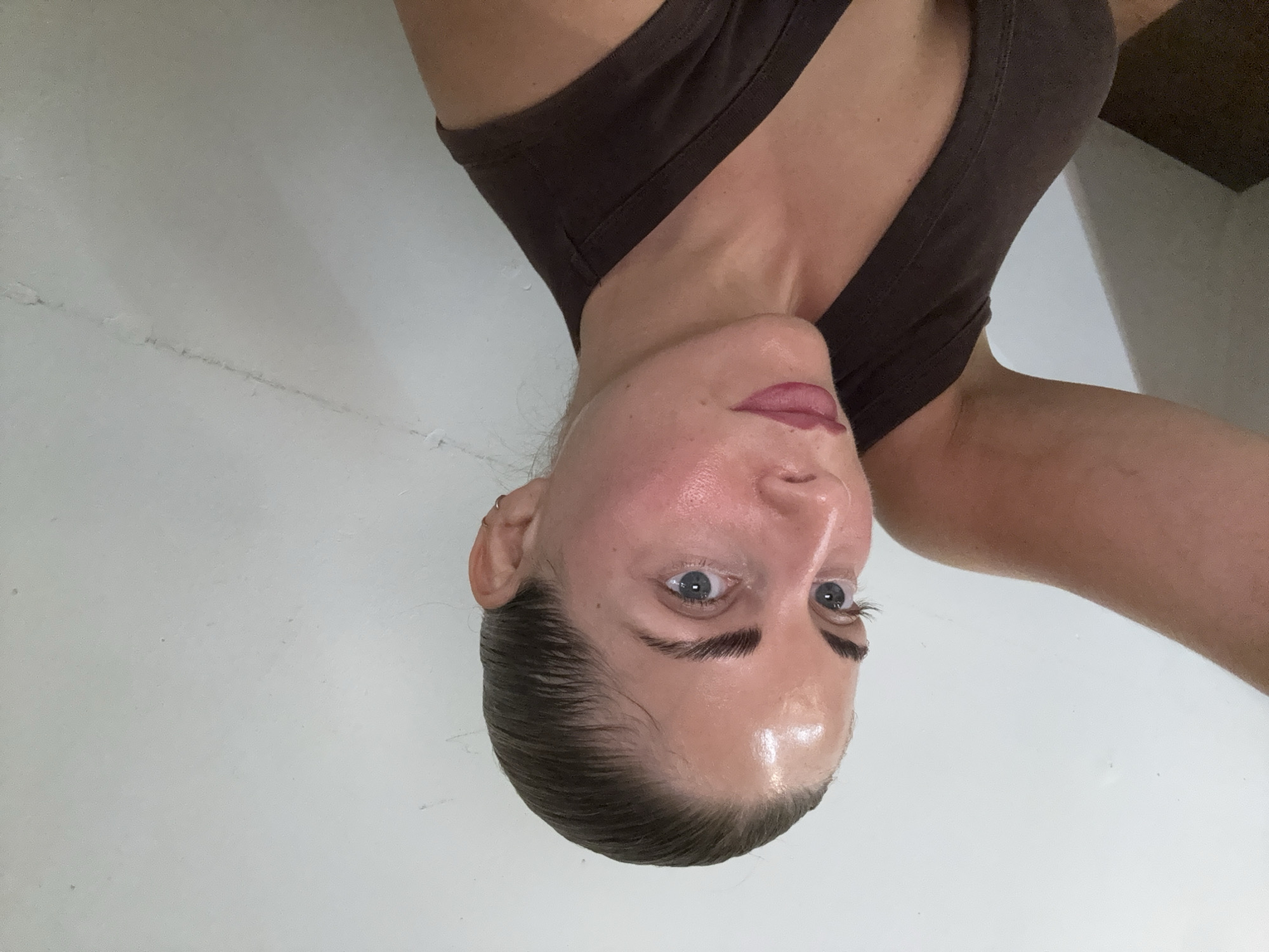 A woman with her hair pulled back is captured in a selfie, her face angled towards the camera. She is wearing a brown, sleeveless top. The background is a plain white wall. Her left arm is raised and extends out of frame. There is no indication of her location in Paris, France.
