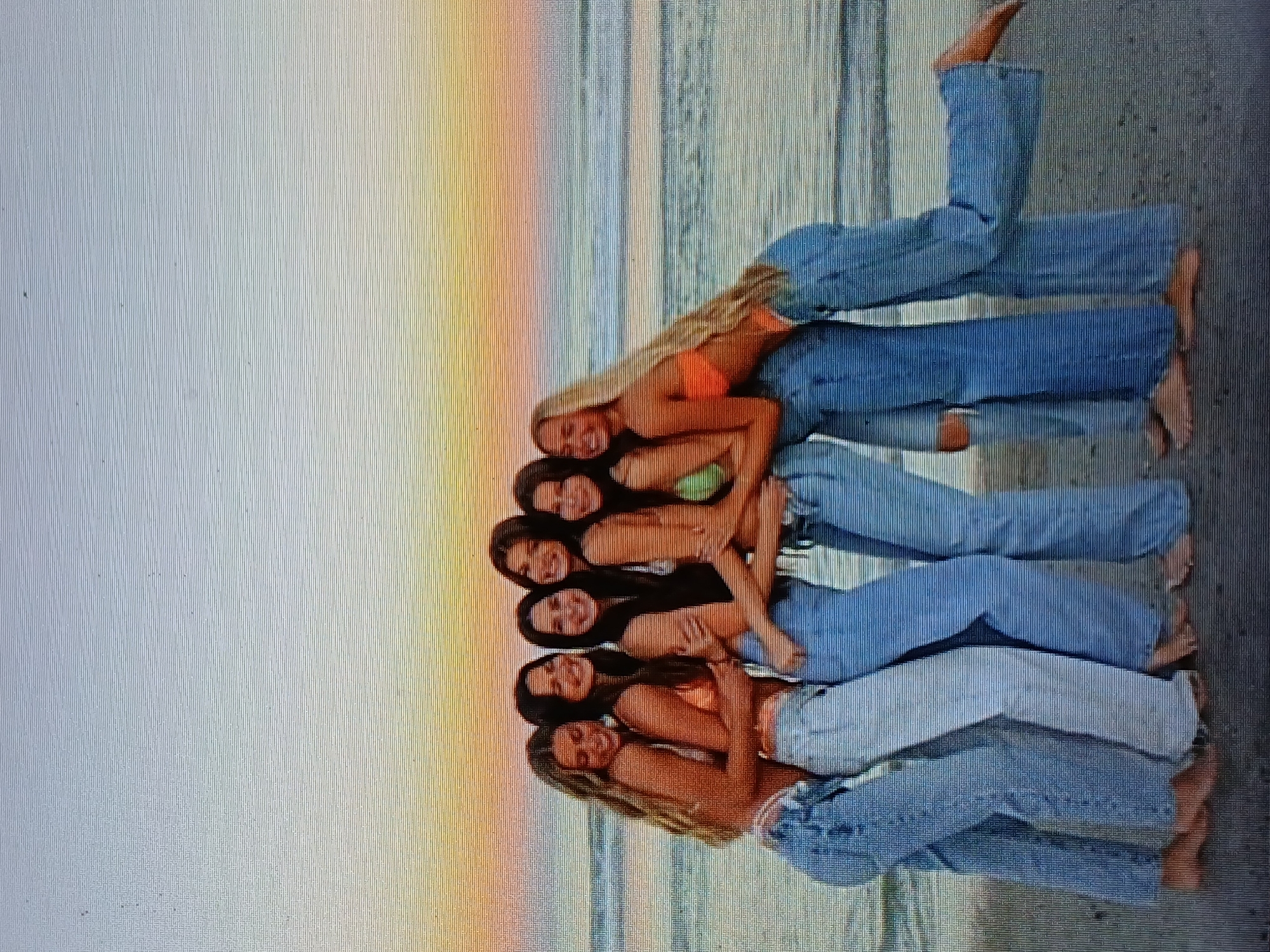 Six young women pose on a beach, likely at sunset. They are lying in a line, one behind the other, with their heads together. All are wearing jeans, and their upper bodies are bare or in swimwear. The background shows the beach and ocean, with a colorful sunset. It is not possible to confirm the specific location as Juan Griego, Venezuela from the image alone.