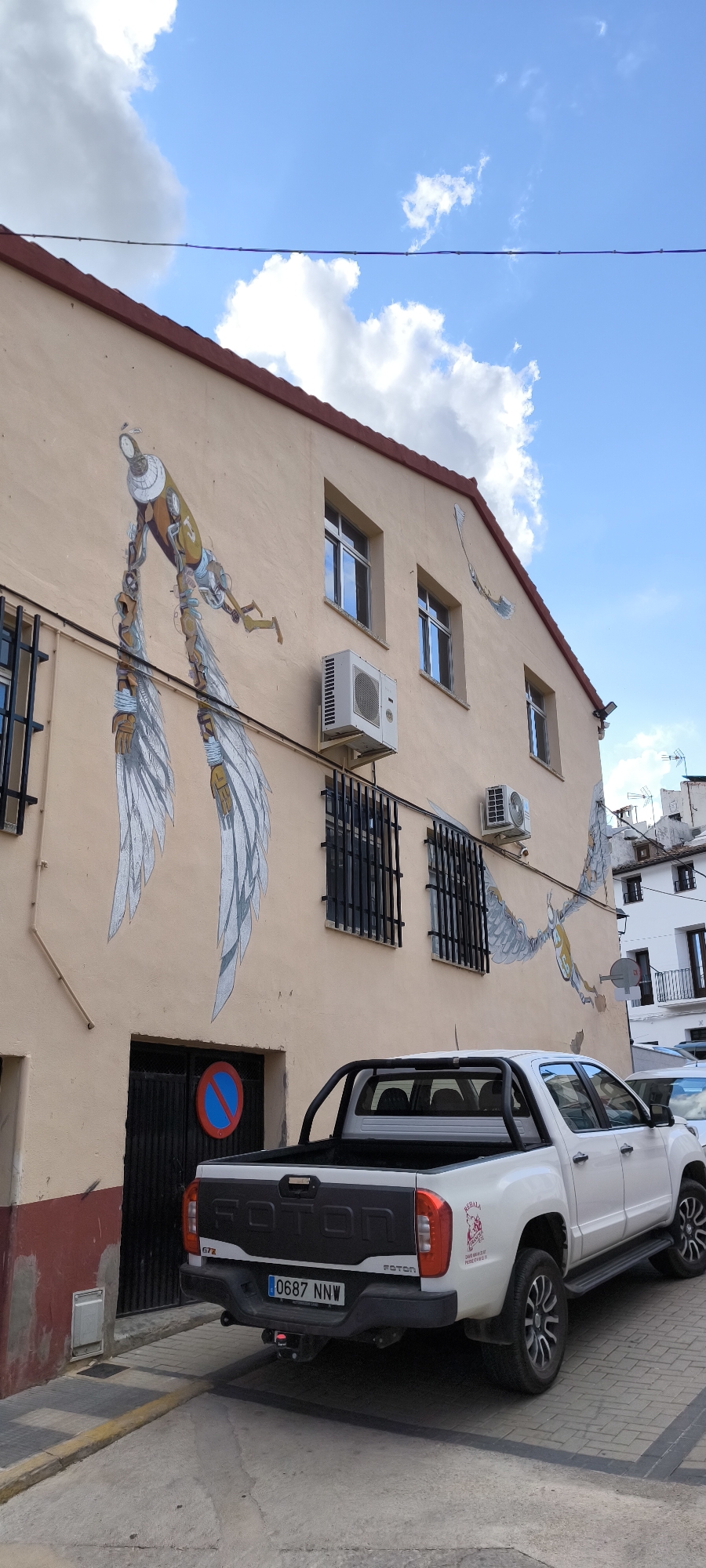 The image shows a street in Fanzara, Spain, with a white pickup truck parked in front of a building. The building has a light-colored facade with murals painted on it, depicting winged figures. There are several windows on the building, some of which are barred. An air conditioning unit is visible on the exterior wall. The truck has a license plate that reads "0687 NNW". The sky is blue with some clouds.