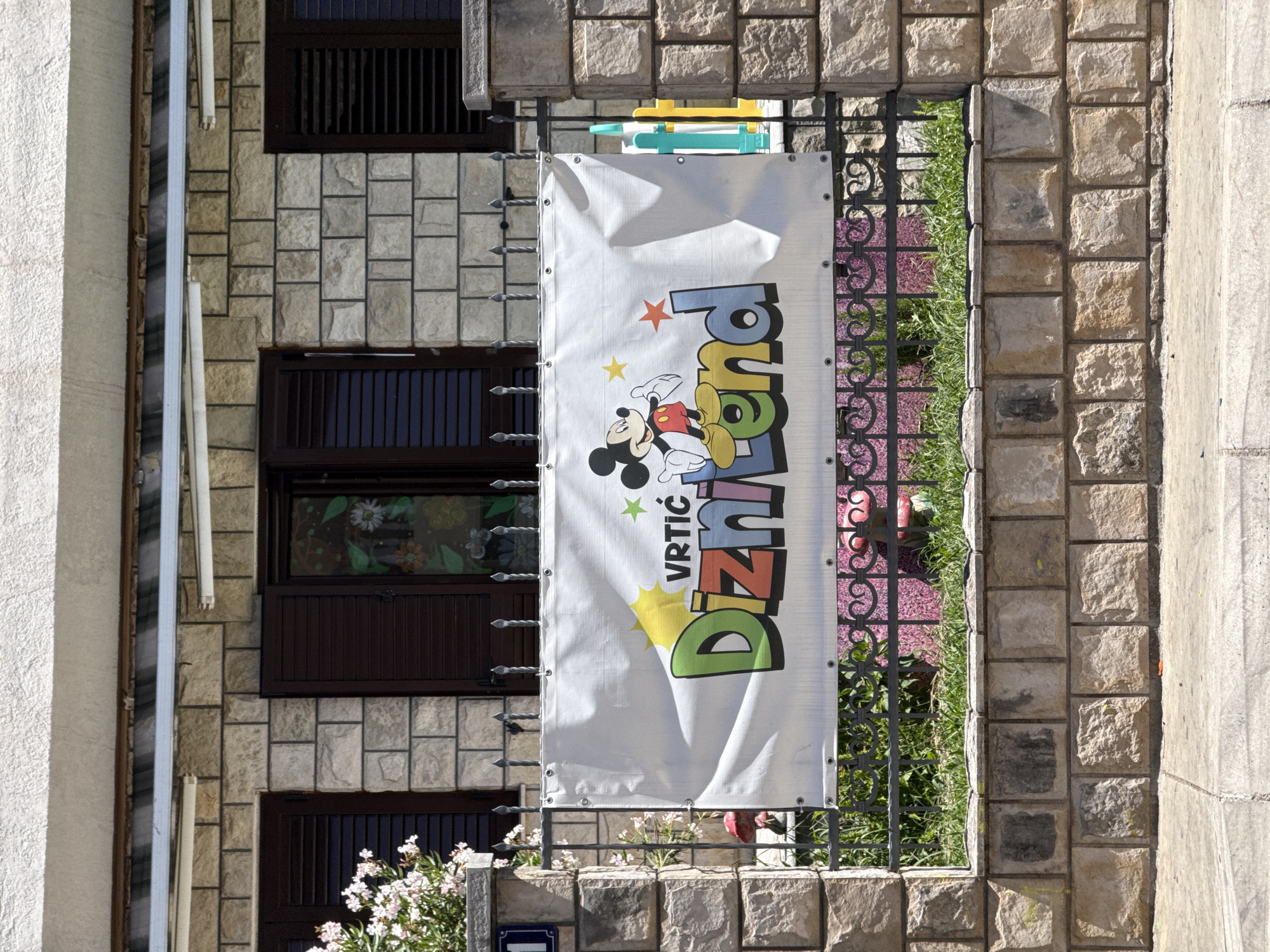 In Budva, Montenegro, a white banner with colorful text and a depiction of Mickey Mouse is displayed on a fence in front of a building. The banner reads "VRTIC Diznilend" and features the iconic Disney character. The building is constructed of stone blocks and has dark-colored shutters on its windows. The fence is made of wrought iron with decorative scrollwork. Behind the fence, green grass and some pink flowers are visible. The area appears to be a street or public walkway, with a concrete sidewalk in the foreground.