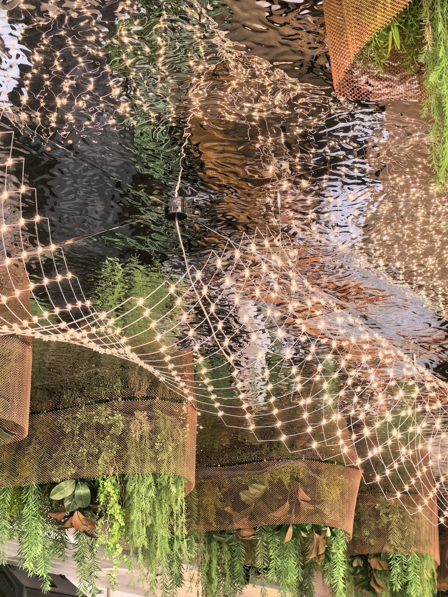 This is a close-up, eye-level shot of a decorative installation featuring a shallow body of water with ripples, reflecting warm-toned string lights. The lights are arranged in a net-like pattern above the water. Below the water's surface, lush green foliage is visible, possibly hanging plants. The overall impression is one of a whimsical, nature-inspired lighting display. The image does not contain any people. There is no information in the image to suggest the location is Repino, Russia.