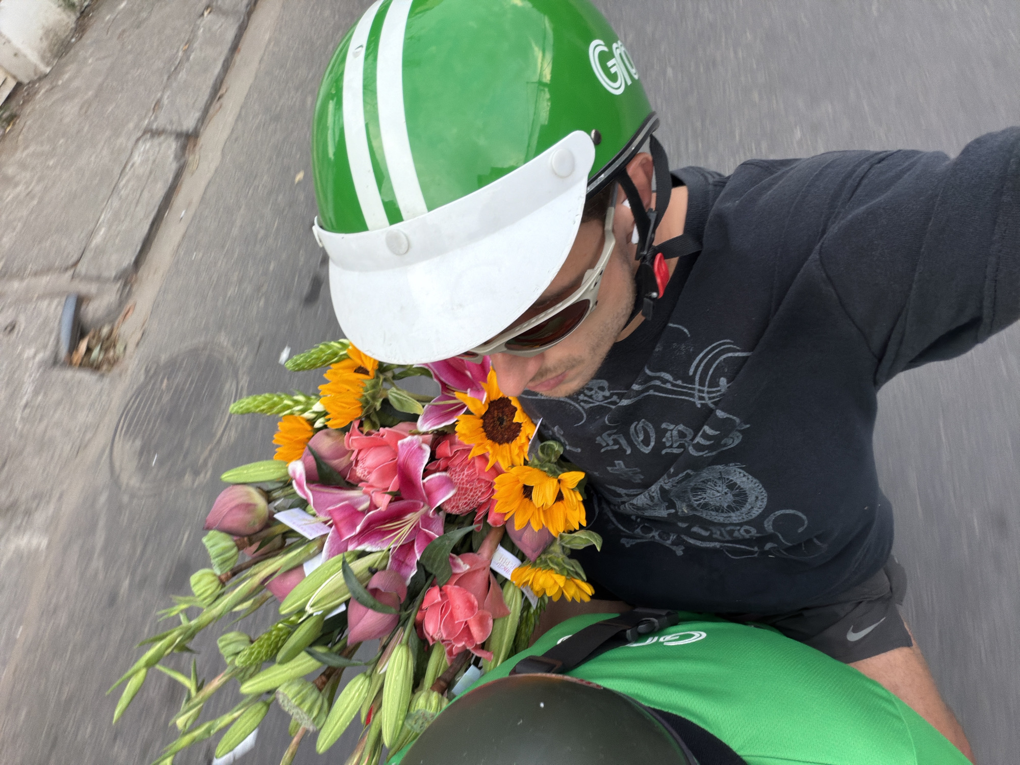 The image presents a high-angle view of a person, likely a delivery rider, transporting a large, vibrant bouquet of flowers on a motorcycle or scooter in what appears to be an urban setting in Da Nang, Vietnam. The perspective suggests a selfie taken while in motion.
The central subject is a male individual wearing a distinctive green helmet with two white vertical stripes and a white visor. The helmet features "Grb" visible on its side, indicative of the "Grab" ride-hailing and delivery service common in Southeast Asia. He is also wearing dark sunglasses and a black long-sleeved t-shirt with a light-colored graphic design. Below, he wears dark shorts with a visible white Nike swoosh logo on the left thigh.
A substantial bouquet of fresh flowers dominates the foreground, held in front of the rider. It comprises bright yellow sunflowers, prominent pink and white lilies, and other pink and reddish-orange blossoms, all surrounded by ample green foliage. The flowers partially obscure the rider's lower face. Beneath the bouquet and the rider's torso, a bright green uniform or delivery bag is visible, with a partial "G" logo, further supporting the Grab affiliation.
The background consists of a grey asphalt road with visible cracks and a curb to the left. The motion blur in the background indicates that the vehicle is moving. The scene takes place during the daytime, under bright but diffused lighting. The overall imagery is consistent with a delivery service worker on duty in a bustling city street, delivering flowers, a common sight in places like Da Nang, Vietnam.