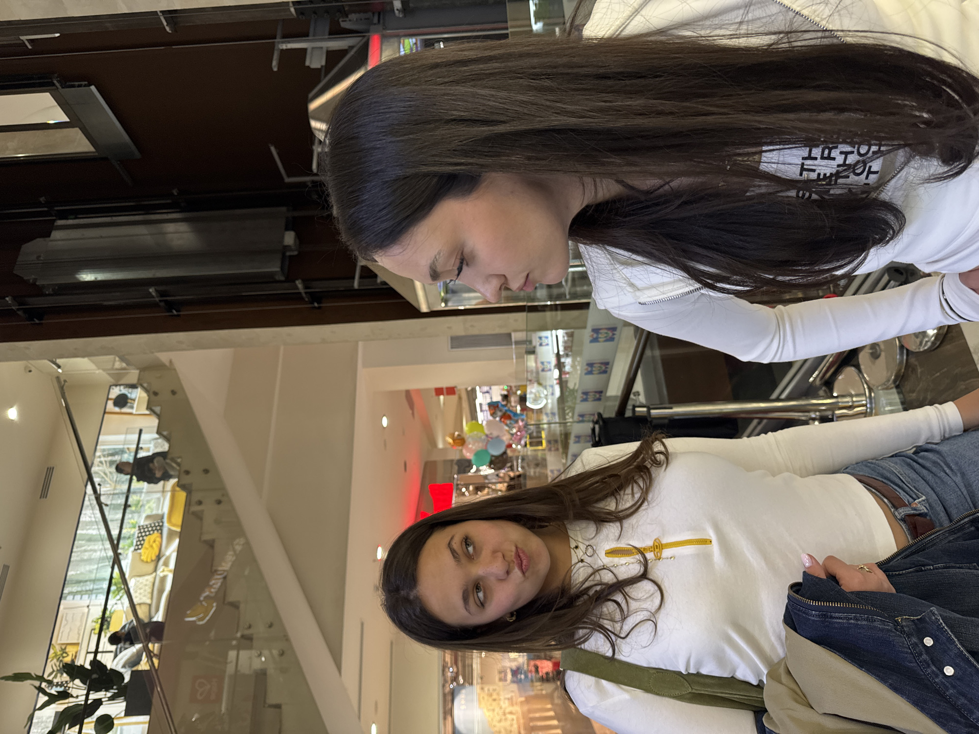 The image depicts two young women inside a modern, multi-level shopping mall. The location appears to be indoors, likely in Yerevan, Armenia, based on the modern architecture and general context.
One woman is wearing a white long-sleeved shirt and blue jeans with a brown belt. She is carrying a green shoulder bag and has a denim jacket draped over her. Her hair is dark and wavy.
The second woman is wearing a white zip-up jacket. Her hair is long, dark and straight.
The background shows elements of a shopping mall interior, including shops, decorative displays, and multiple levels. There is a glass partition visible, suggesting an elevated viewpoint. The environment indicates a commercial, urban setting.