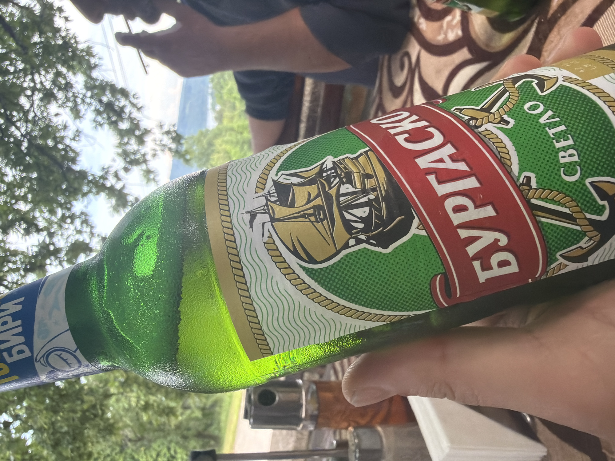 This image depicts a casual outdoor scene in Sushina, Bulgaria, under bright daylight. In the foreground, a hand holds a cold, green glass bottle of beer, evidenced by the condensation on its surface.
The bottle's label is prominent, featuring Cyrillic script. A red band displays "БУРГАСКО" (Burgasko), identifying the brand. Below this, on a green background, the word "СВЕТЛО" (Svetlo), meaning "light," indicates the beer type. The label also showcases a detailed illustration of a sailing ship, surrounded by anchors and rope patterns. On the bottle's neck, "БИРИ" (Biri), plural for beer, is visible.
In the upper left background, a person is partially visible, seen from the chest up, seemingly relaxed. They are holding a thin object, possibly a cigarette or a straw, to their mouth. The backdrop is filled with lush green trees and foliage, suggesting a natural and verdant environment. Beyond the immediate trees, a hazy, distant landscape, likely mountains or hills, is visible under a bright sky, reinforcing the outdoor setting. The presence of a patterned tablecloth under the bottle hints at a table, suggesting a relaxed gathering on a patio, balcony, or outdoor eating area. The overall mood is calm and informal.
