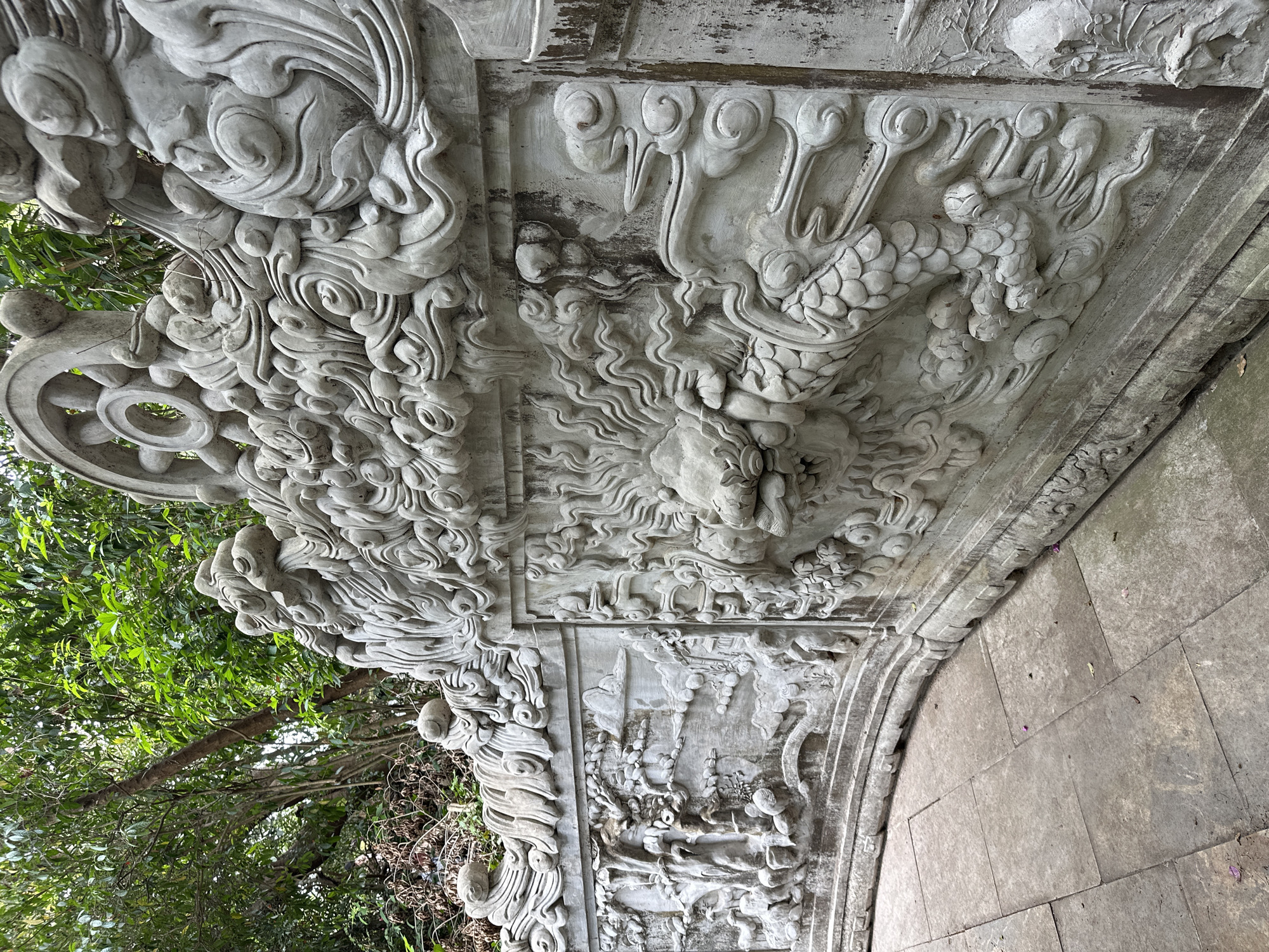 The image shows a close-up of an ornate, curved stone balustrade featuring intricate bas-relief carvings of mythical creatures, possibly dragons and clouds, with some floral motifs. The stone is weathered and shows signs of age and possibly some staining. In the background, lush green foliage from trees is visible. The balustrade appears to be part of an outdoor architectural feature, possibly in a garden or temple setting. The paving stones in the foreground are large, rectangular, and laid in a grid pattern. There are no people or animals visible in the image. This balustrade is located at the Son Tra Peninsula in Da Nang, Vietnam, specifically within the Linh Ung Pagoda complex.