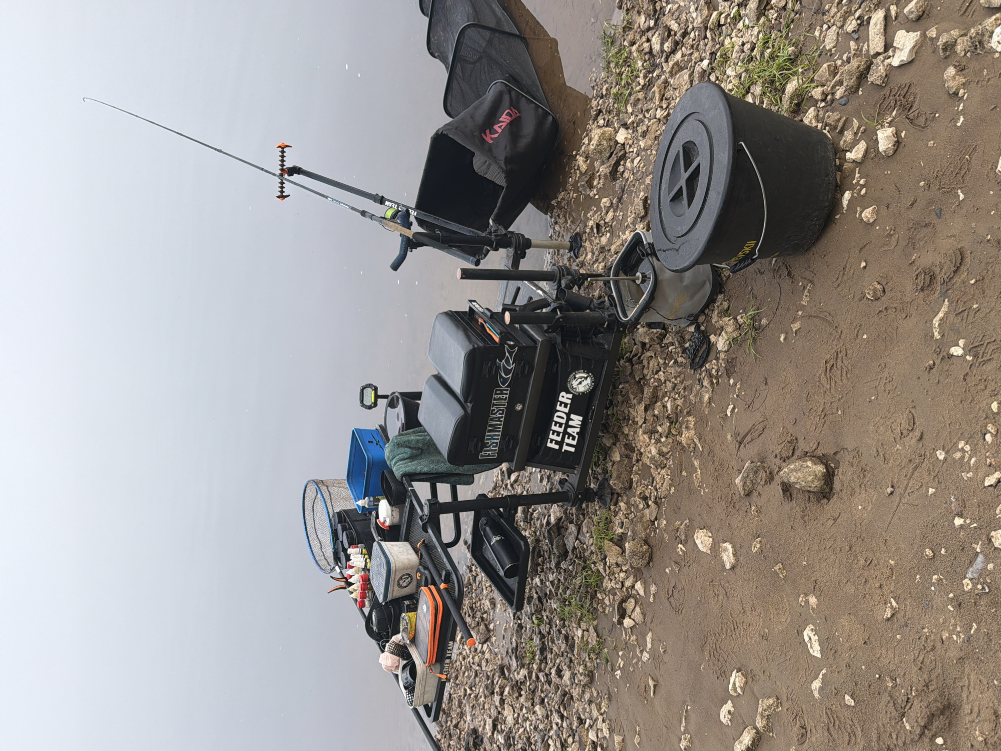 The image captures a comprehensive feeder fishing setup positioned on the bank of a body of water, likely a river or large lake, in Noviy, Russia. The bank consists of a blend of small rocks and wet, sandy soil. The water surface is calm and appears murky, reflecting an overcast, uniform grey sky. No individuals are visible in the scene.
A black fishing seat box, labeled "FISHMASTER" and "FEEDER TEAM," serves as the central station, equipped with various attachments. One fishing rod extends over the water, its tip visible against the grey backdrop. A black keepnet, bearing the red text "KAIDA," is partially submerged next to the station. On the sandy bank, a large black lidded bucket is visible. The fishing station is further accessorized with numerous smaller containers, tackle boxes, a blue container, a small landing net, and a digital counter, all meticulously arranged for a fishing session.
The arrangement of rods, the ready keepnet, and the organized tackle suggest an active or ongoing feeder fishing activity. The overcast sky indicates daytime conditions. The wet ground and murky water are consistent with a river or lake environment, portraying a scene of dedicated angling preparation.