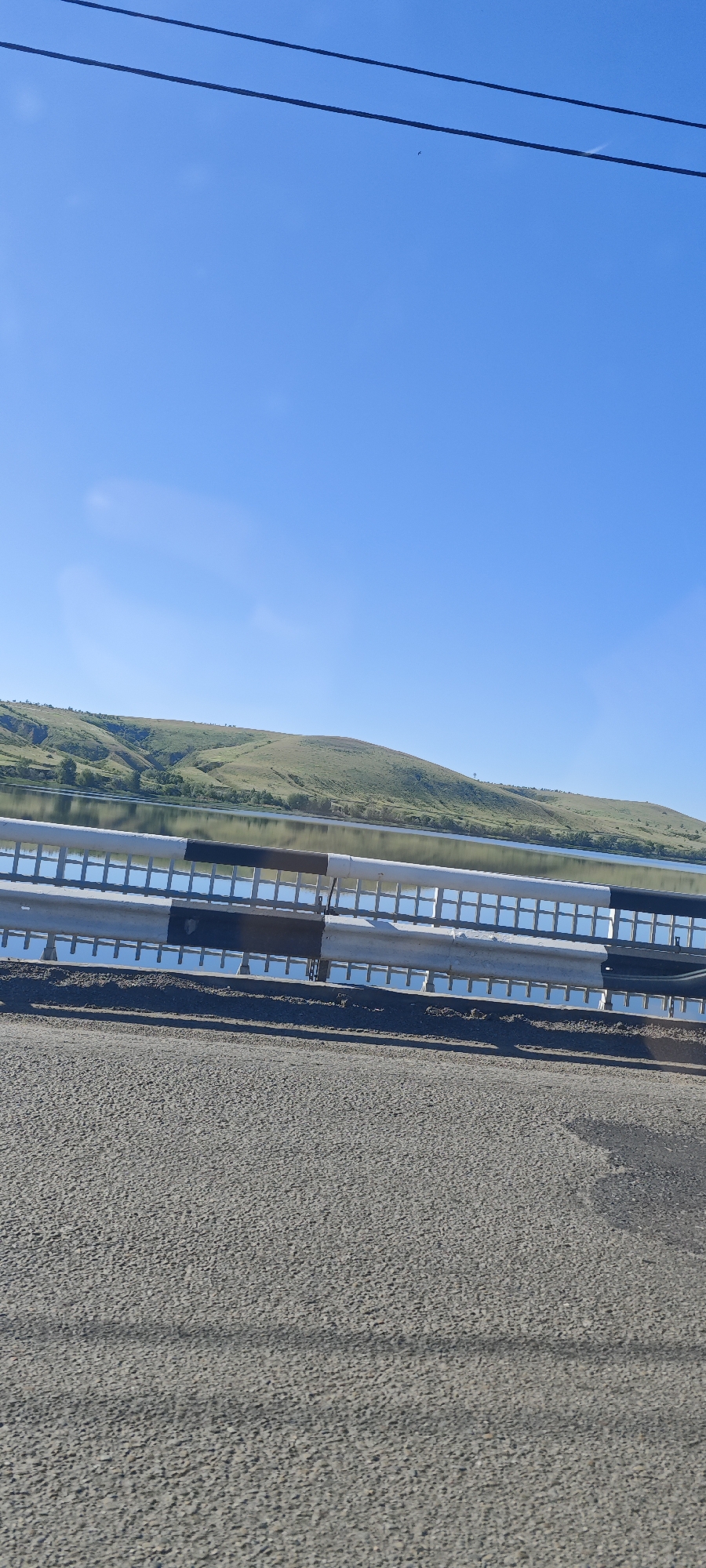 The image shows a view from a bridge in Ferma Sovkhoz Balykleyskiy, Russia. A gray asphalt road is in the foreground. A metal guardrail separates the road from the water. Beyond the guardrail is a body of water, likely a river or lake, reflecting the sky. Green, grassy hills rise from the opposite bank of the water. The sky is clear and blue. Two power lines extend across the top of the image.