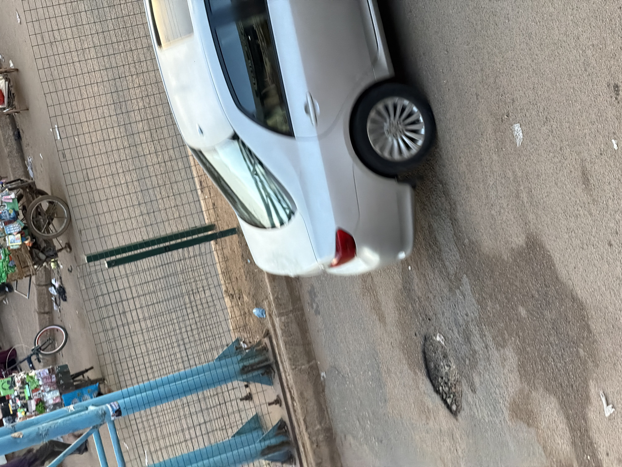 A silver sedan is parked on a paved road. The car is facing away from the camera and towards the right side of the frame. In the background, there are several market stalls and people. The market stalls are loaded with goods. There is a bicycle parked near one of the stalls. The scene appears to be in a busy urban environment. The road is cracked and uneven, with a large pothole visible in the foreground. There is also some debris scattered on the road.