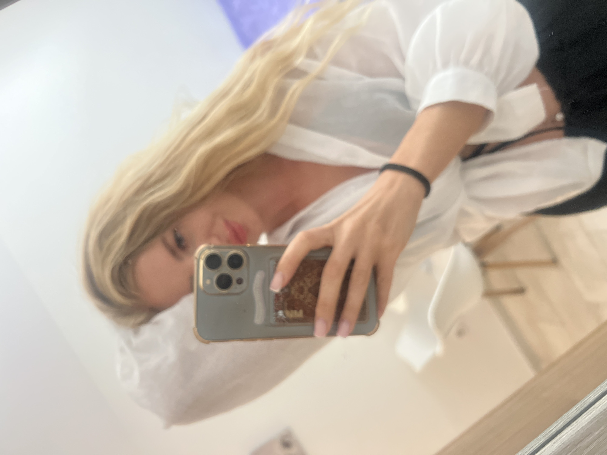 This is a mirror selfie taken indoors in Budva, Montenegro, featuring a woman with long, wavy blonde hair. She appears to be lying down or reclining, with her head resting on a light-colored, possibly white, fabric object.
The woman is wearing a loose-fitting white blouse or shirt, under which a black bra strap is visible. A black hair tie is worn on her left wrist. She is holding a smartphone, identifiable by its multi-lens camera system and gold-rimmed case. The back of the phone case has a textured, possibly patterned, design. Her fingers, with light pink manicured nails, are visible around the phone. Her face is partially captured in the reflection, showing a neutral to slightly relaxed expression with slightly parted lips.
The image is captured through a large reflective surface, likely a mirror or polished table, which also shows a blurry reflection of a room with light-colored walls and some wooden furniture or framing along the bottom right. The bright lighting suggests it is daytime. No discernible text is visible in the image.