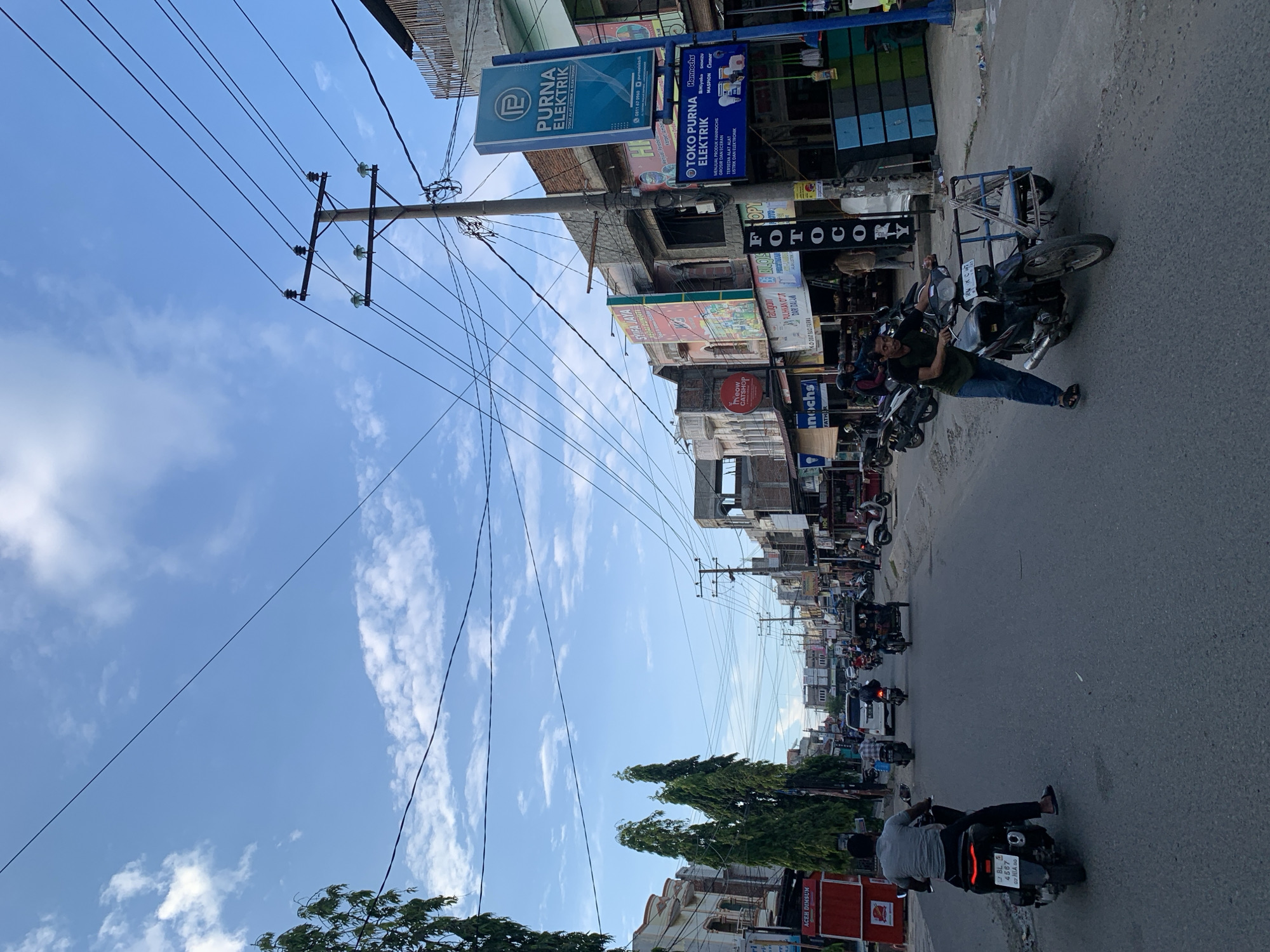 A street scene in Lhokseumawe, Indonesia, is depicted on a partly cloudy day. The road is paved and appears to be a main thoroughfare with a significant amount of traffic. Several motorcycles, including one with a sidecar, are visible, along with an auto-rickshaw and a van. Pedestrians are also present, walking on the sidewalks and in the street.
The buildings lining the street are varied in architecture and condition. Some are multi-story commercial buildings with awnings and signage in Indonesian, while others appear to be simpler structures. Prominent signs include "Purna Elektrik" and "Toko Purna Elektrik," indicating electrical supply stores. There are also signs for "Aceh Dimsun" and what appears to be a cat-themed establishment.
The environment features tall, slender trees planted along the roadside, and a complex network of power lines overhead supported by utility poles. The sky is a bright blue with scattered white clouds. The overall impression is of a bustling urban environment with active commerce and daily life taking place on the street. The light suggests it is likely late afternoon or early evening.